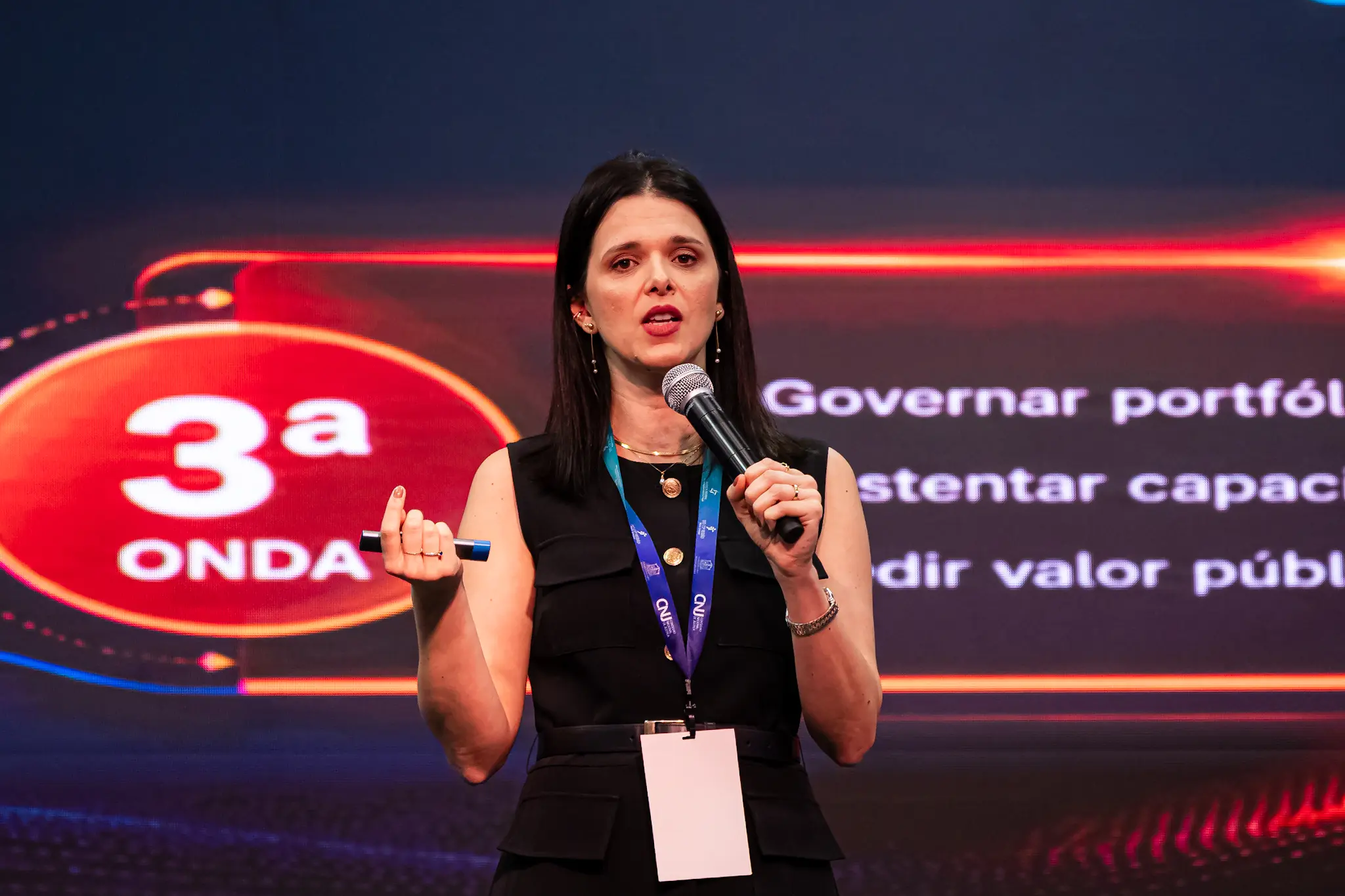 Plano médio de uma mulher de cabelos escuros e vestido preto, usando crachá. Ela segura um microfone próximo à boca com a mão esquerda e um passador de slides na mão direita. Ao fundo, um painel exibe o texto "3ª ONDA".