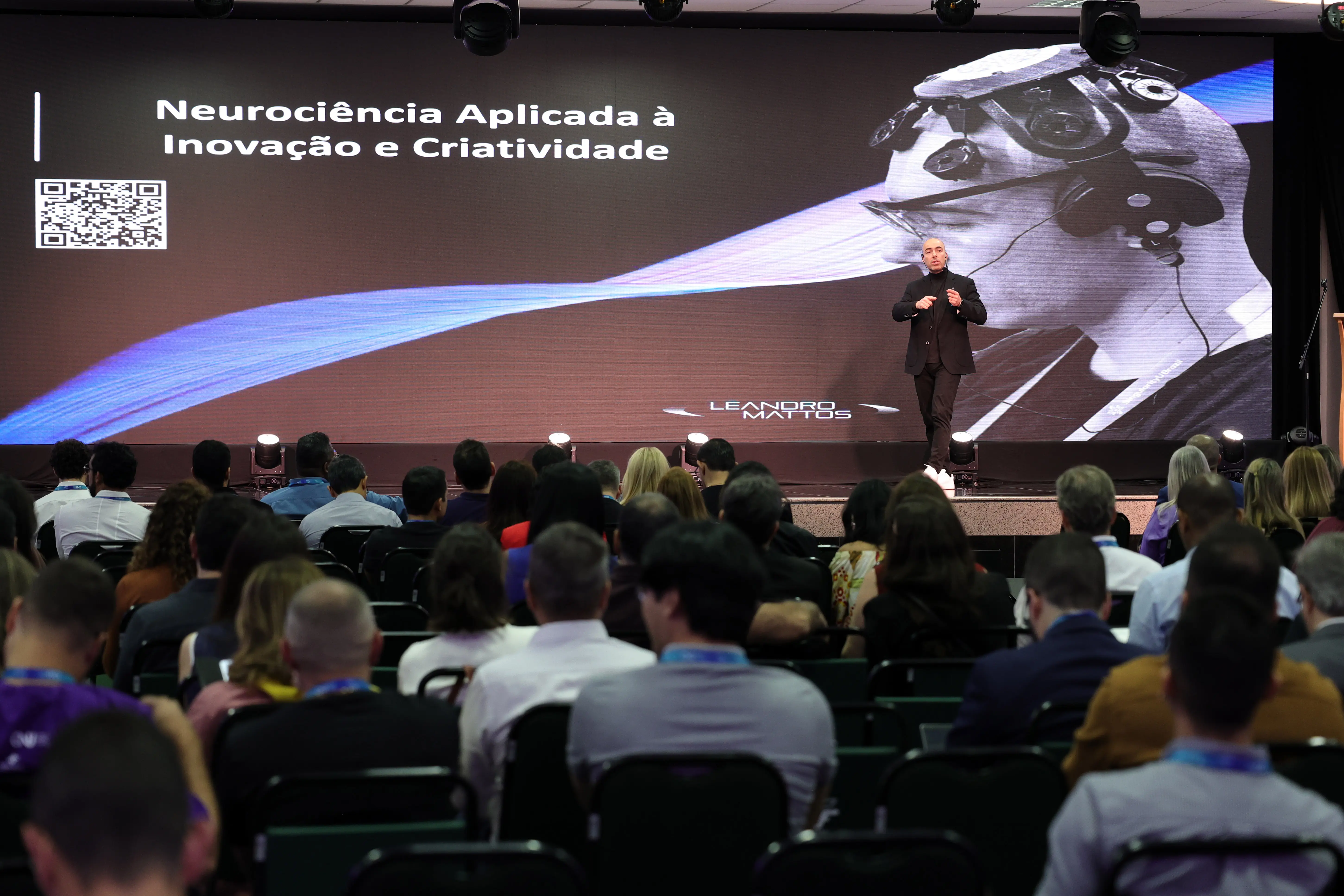 Visão geral de uma palestra em auditório. No palco, um palestrante de preto apresenta-se diante de um telão com o título "Neurociência Aplicada à Inovação e Criatividade" e a foto em preto e branco de um perfil usando um dispositivo na cabeça.