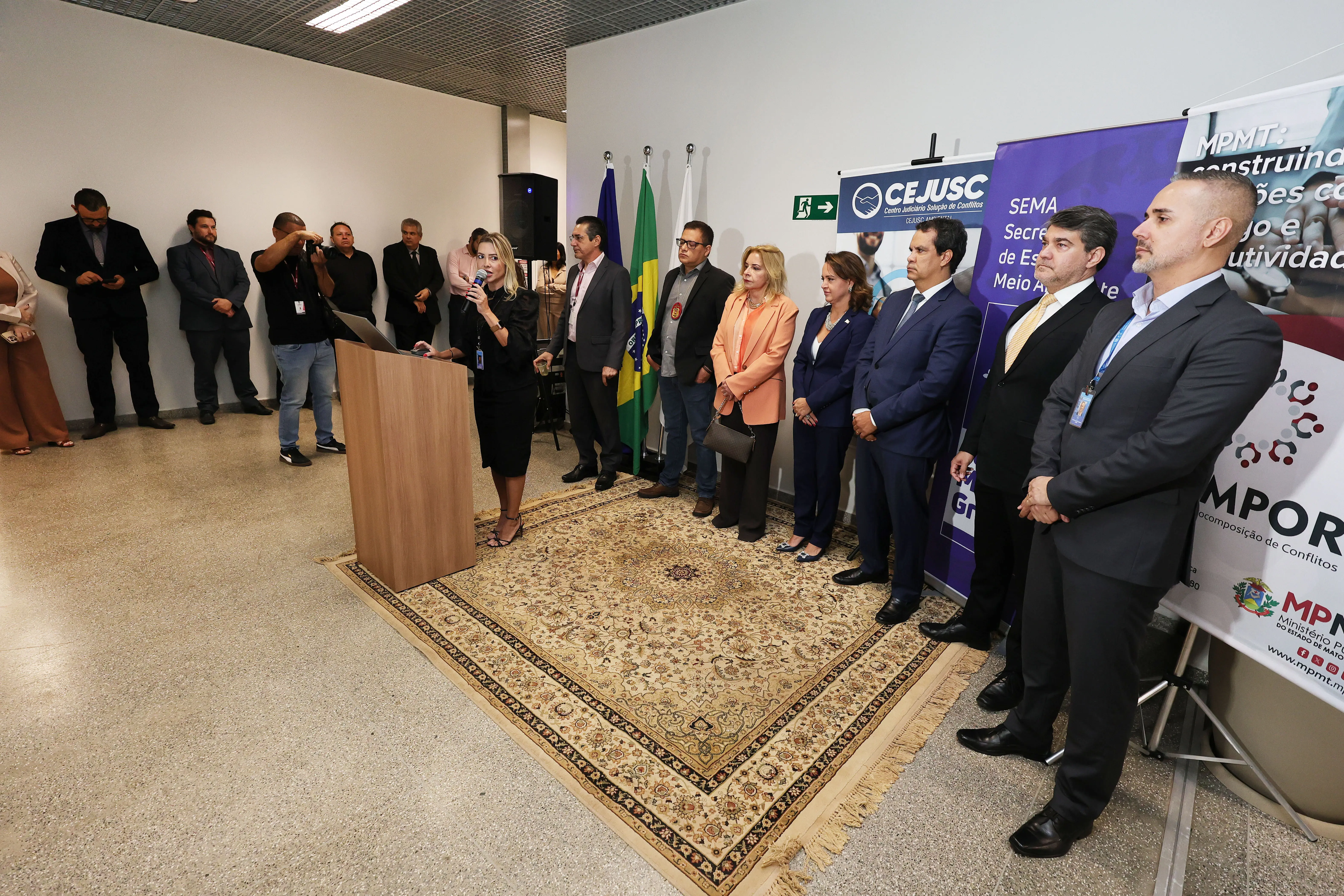 Uma mulher fala ao microfone em um púlpito de madeira durante evento institucional. À sua direita, autoridades de terno e blazer estão perfiladas sobre um tapete estampado. Ao fundo, há bandeiras e banners institucionais do CEJUSC, SEMA e MPMT.