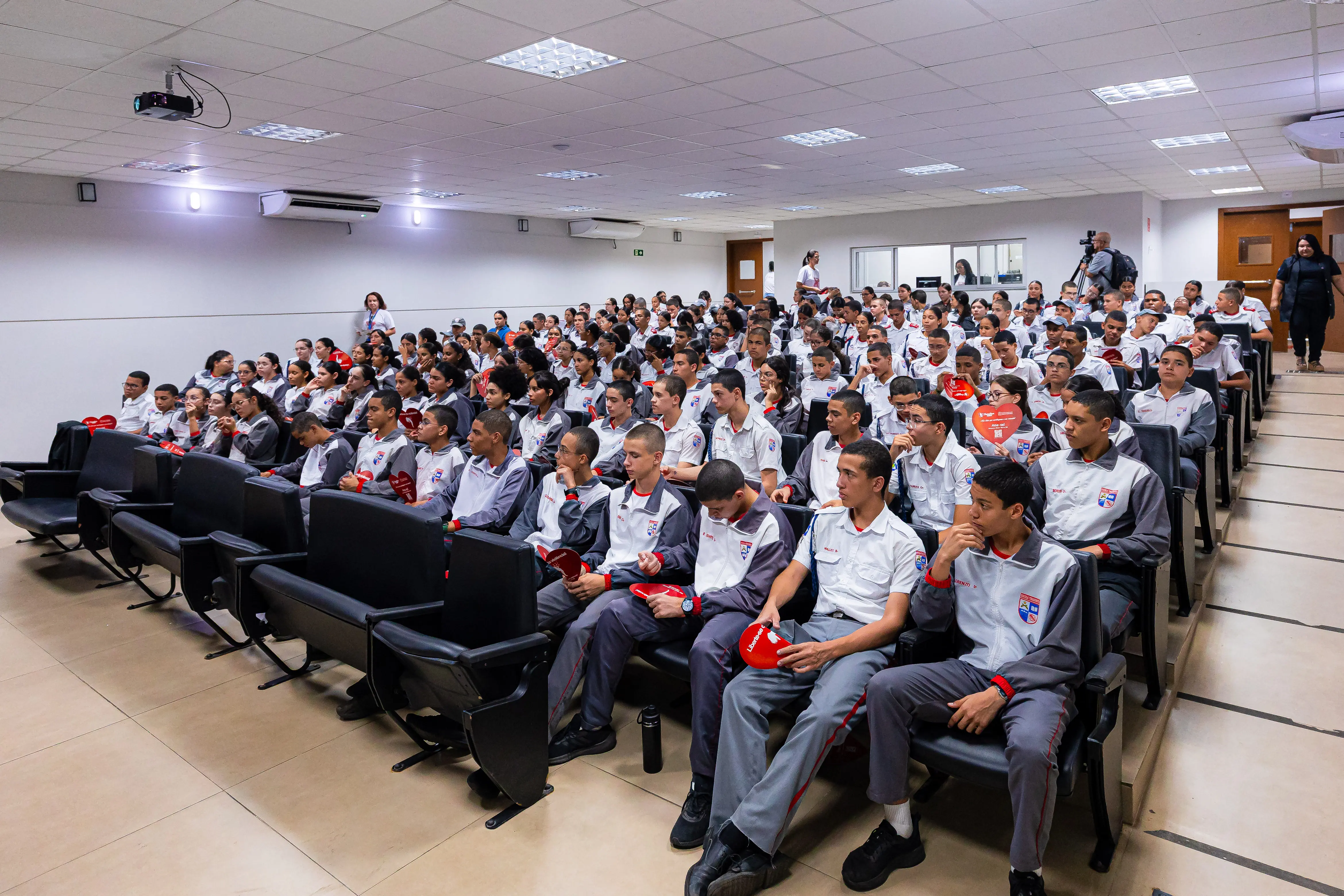 Estudantes uniformizados, em jaquetas cinza e branca, sentados em auditório lotado, seguram corações vermelhos e assistem atentamente à palestra sobre violência doméstica.