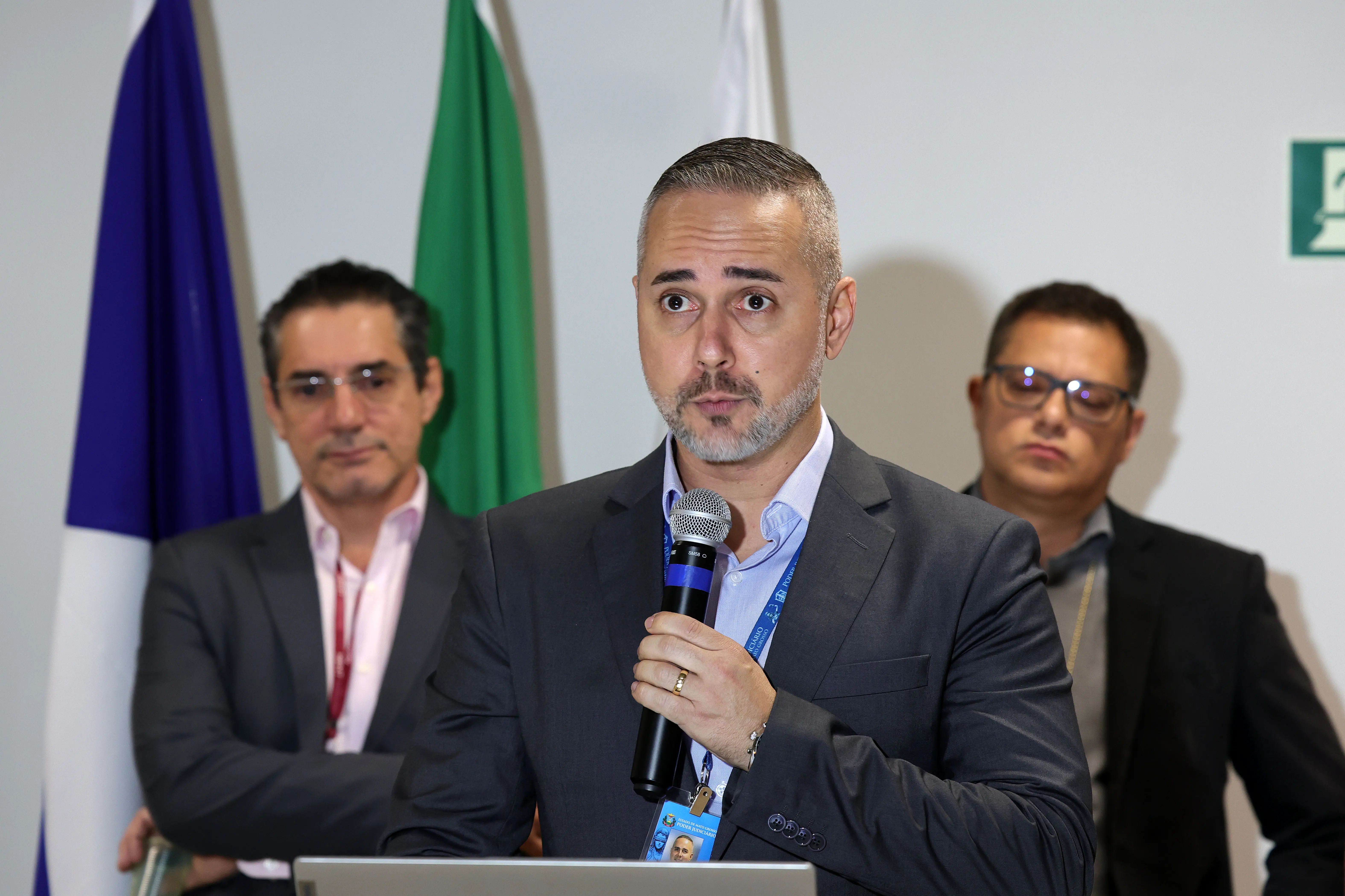 Um homem de terno cinza-escuro e barba grisalha fala ao microfone em primeiro plano. Ele segura o microfone com a mão direita e usa crachá de identificação. Ao fundo, desfocados, dois homens de terno observam a fala diante das bandeiras institucionais.