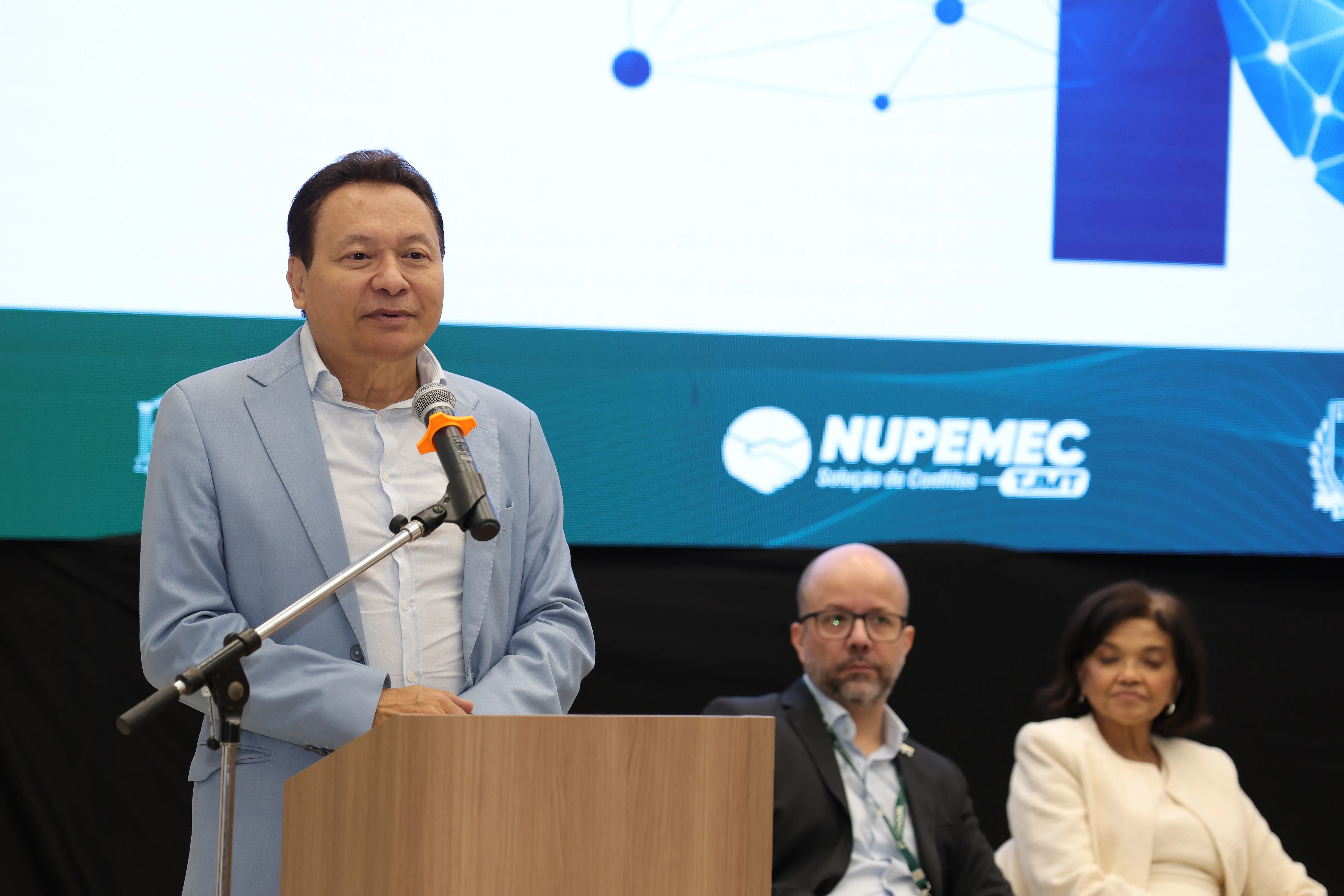 Homem de terno azul claro discursa em um púlpito de madeira com microfone acoplado. Ele olha para a esquerda e sorri levemente. Ao fundo, uma tela projeta logotipos brancos sobre fundo azul, incluindo "NUPEMEC". Um homem e uma mulher estão sentados atrás dele.