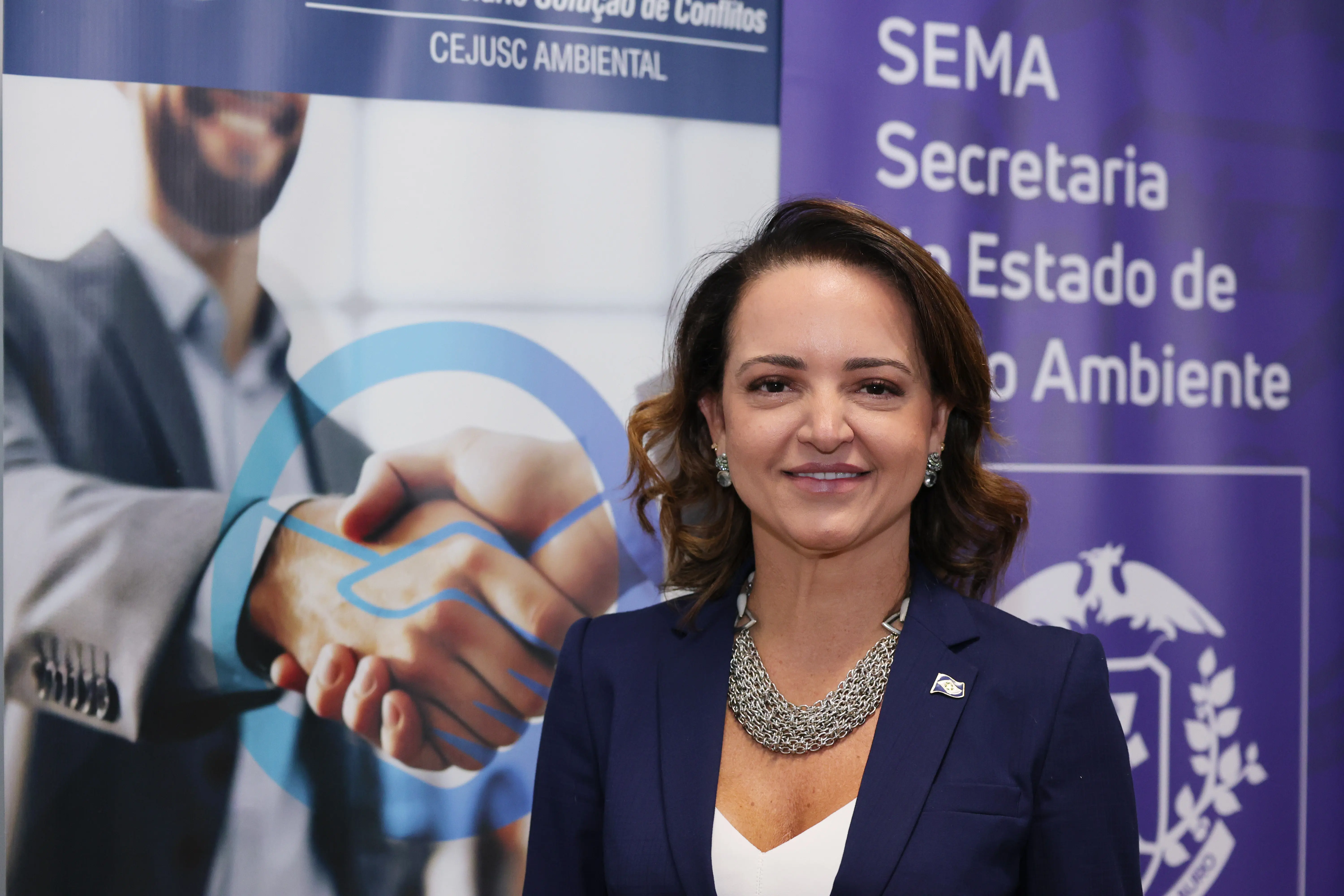 Retrato em plano médio de uma mulher sorrindo, vestindo blazer azul-marinho, blusa branca e colar prateado largo. Ela está posicionada à frente de um banner com ilustrações de aperto de mãos e a inscrição "SEMA Secretaria do Estado de Meio Ambiente".