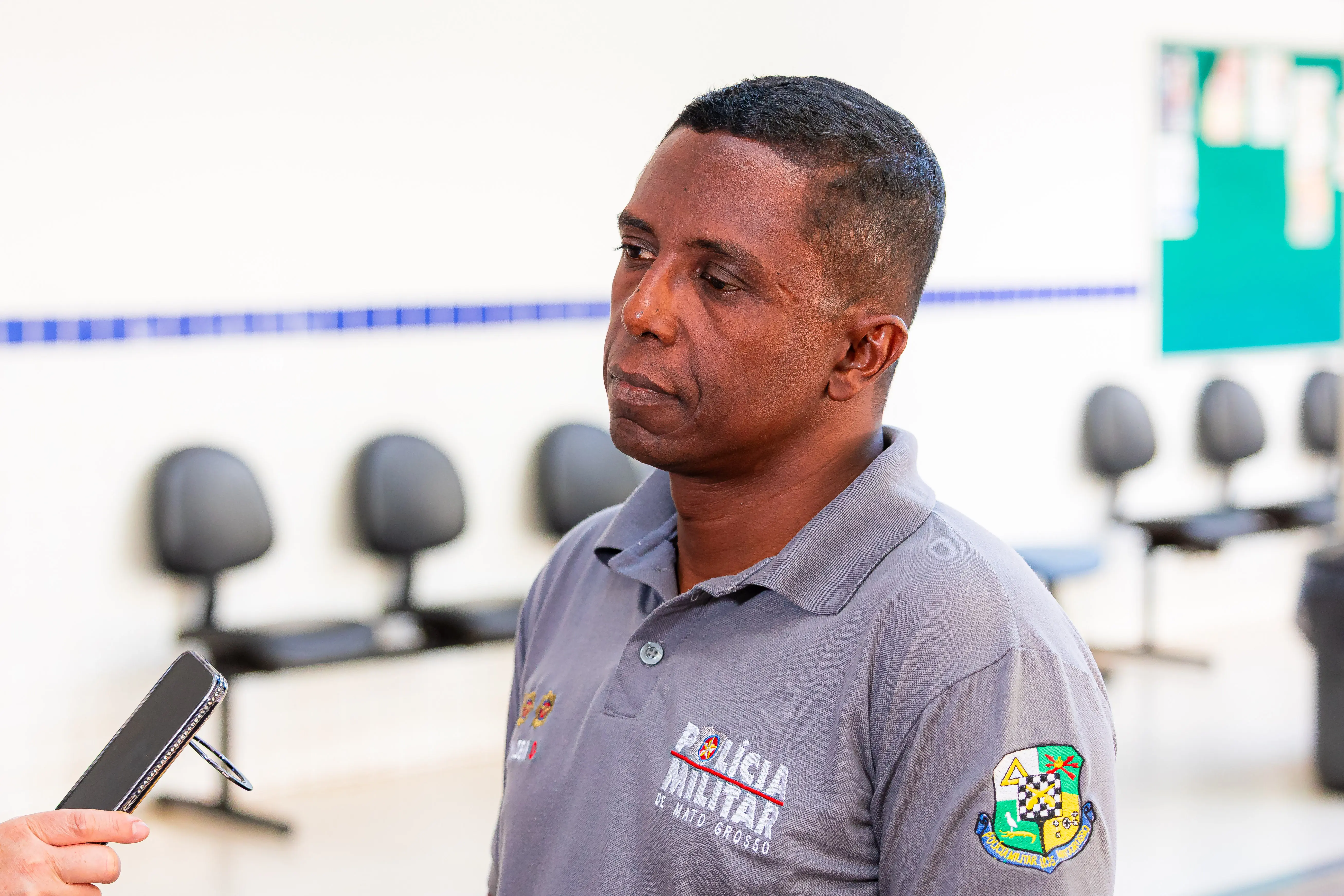 Homem de farda cinza da Polícia Militar de Mato Grosso é entrevistado com microfone de celular, semblante sério, em sala com cadeiras ao fundo.
