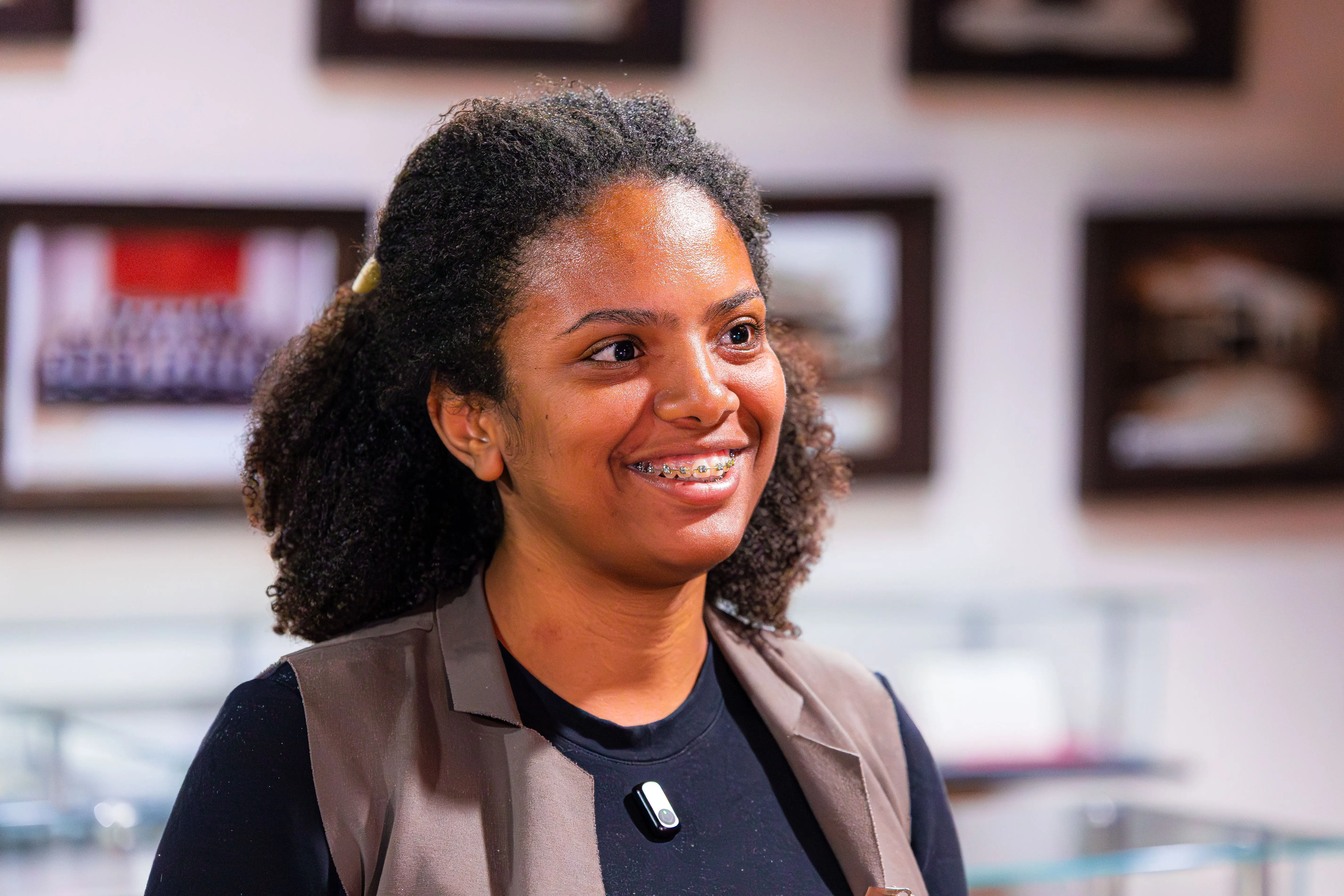 Mulher negra de cabelo cacheado preso para trás sorri usando aparelho nos dentes. Veste blusa preta, colete cinza e lapela. Ao fundo, uma parede com quadros emoldurados desfocados.