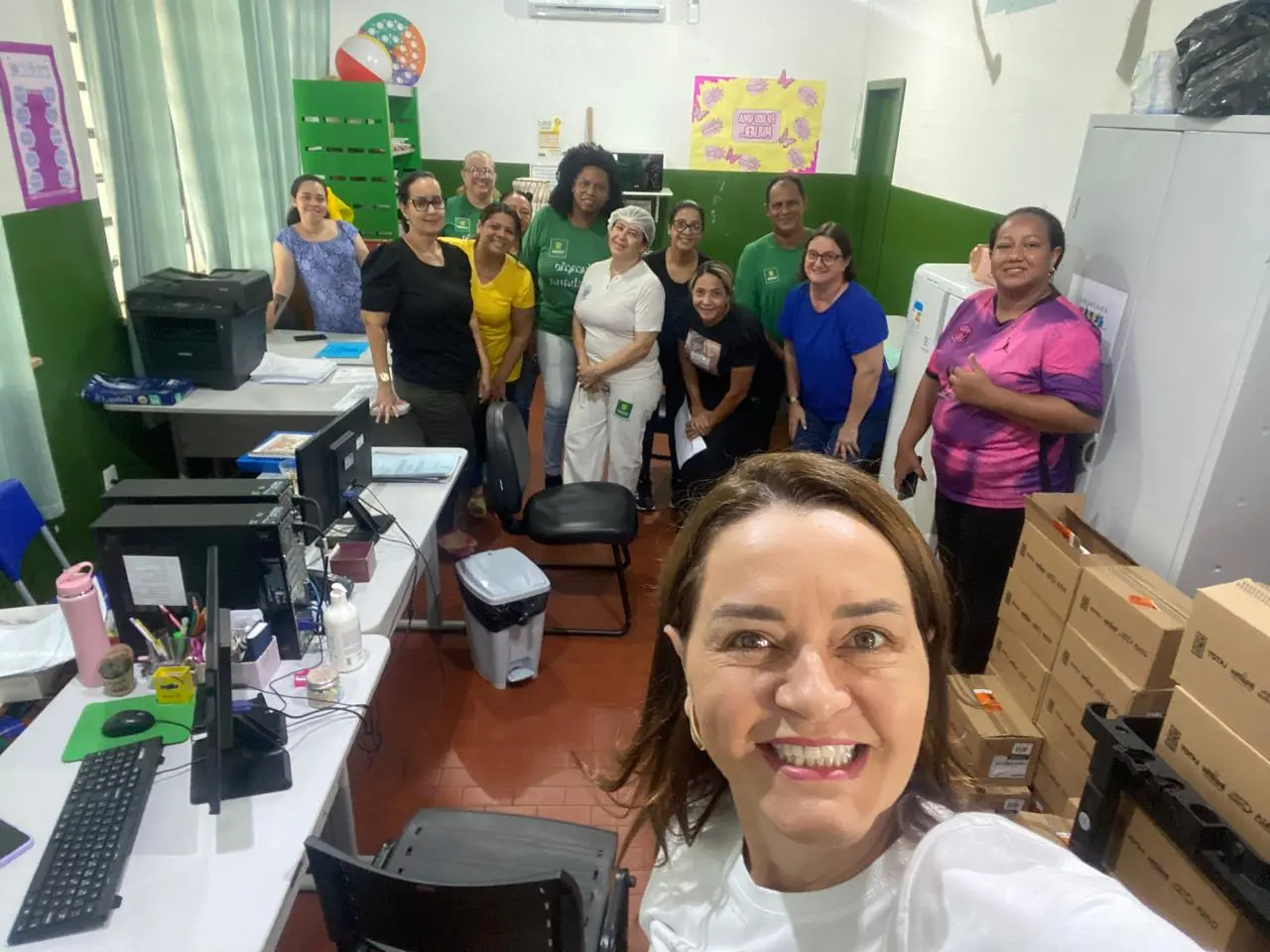 A imagem é uma fotografia em formato selfie tirada na secretaria da escola. Em primeiro plano, no canto inferior direito, uma mulher de cabelos castanhos claros na altura dos ombros e sorriso amplo segura a câmera. Ao fundo, um grupo de cerca de dez pessoas (homens e mulheres) está reunido e sorrind
