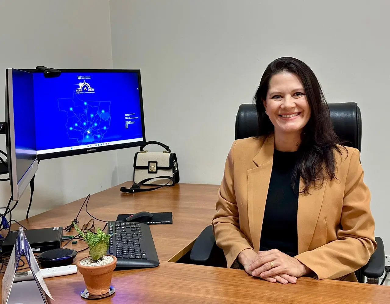 Mulher de cabelos escuros e sorriso amigável, veste blazer caramelo e blusa preta, sentada à mesa de escritório de madeira. Suas mãos estão cruzadas sobre a mesa. Ao fundo, dois monitores de computador exibem o mapa de Mato Grosso. 