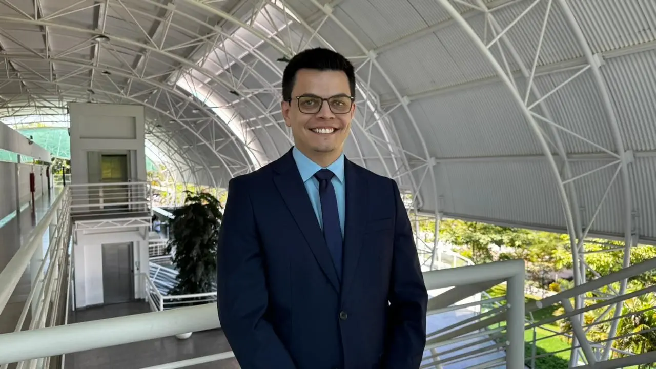 Fotografia em plano médio de um homem sorrindo, vestindo terno azul marinho, camisa azul claro, gravata azul marinho e óculos. Ele está em uma passarela com estrutura moderna, telhado em arco e treliças brancas. 