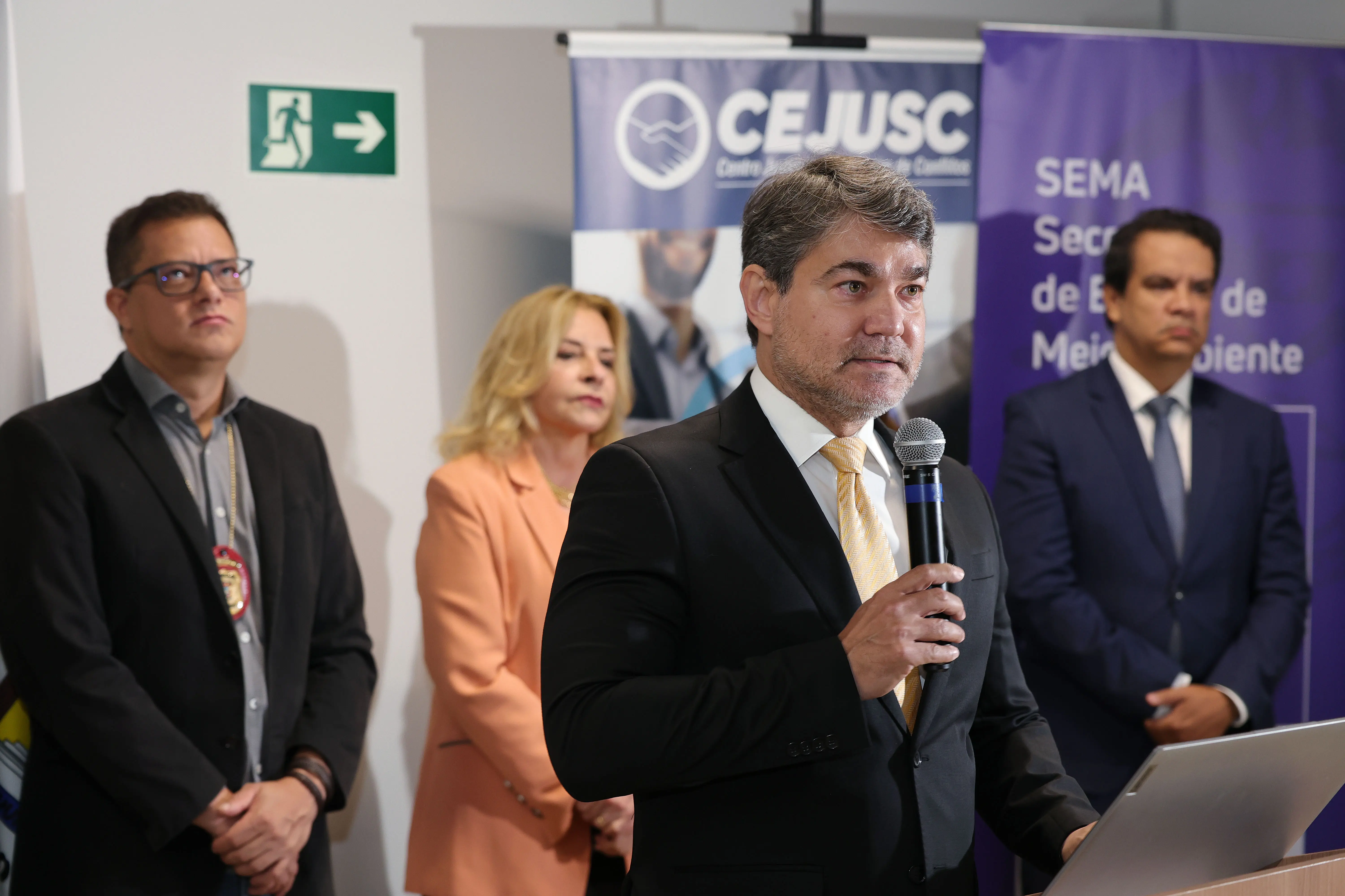 Um homem de terno preto e gravata amarela fala ao microfone em primeiro plano, posicionado atrás de um púlpito com notebook. Ao fundo, em plano intermediário, três pessoas de traje formal acompanham a apresentação em frente a banners do CEJUSC e da SEMA.
