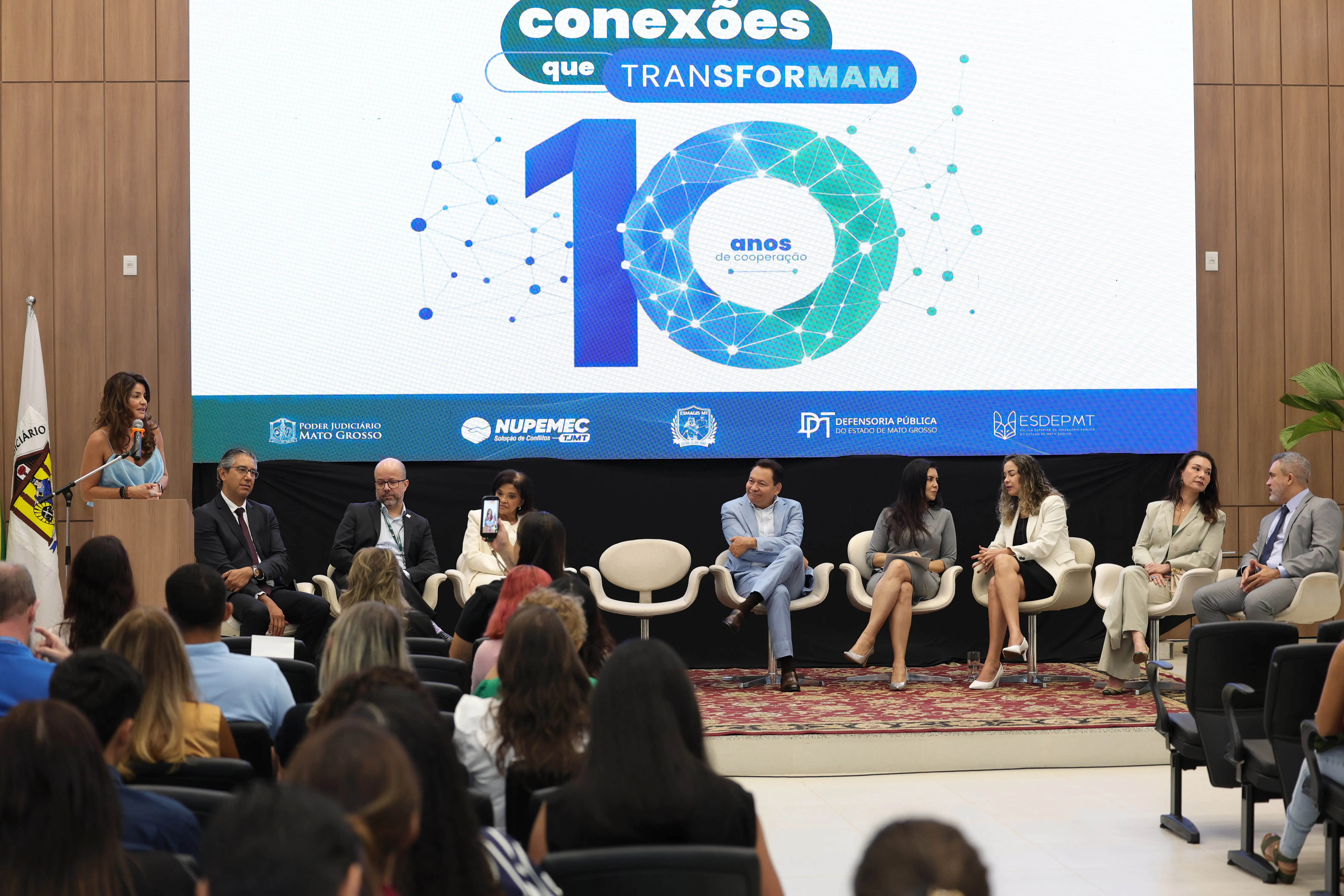 Visão geral do auditório com plateia de costas em primeiro plano. No palco, uma mulher fala ao microfone no púlpito à esquerda, enquanto oito pessoas em trajes formais estão sentadas em poltronas brancas sob um grande painel azul e branco.
