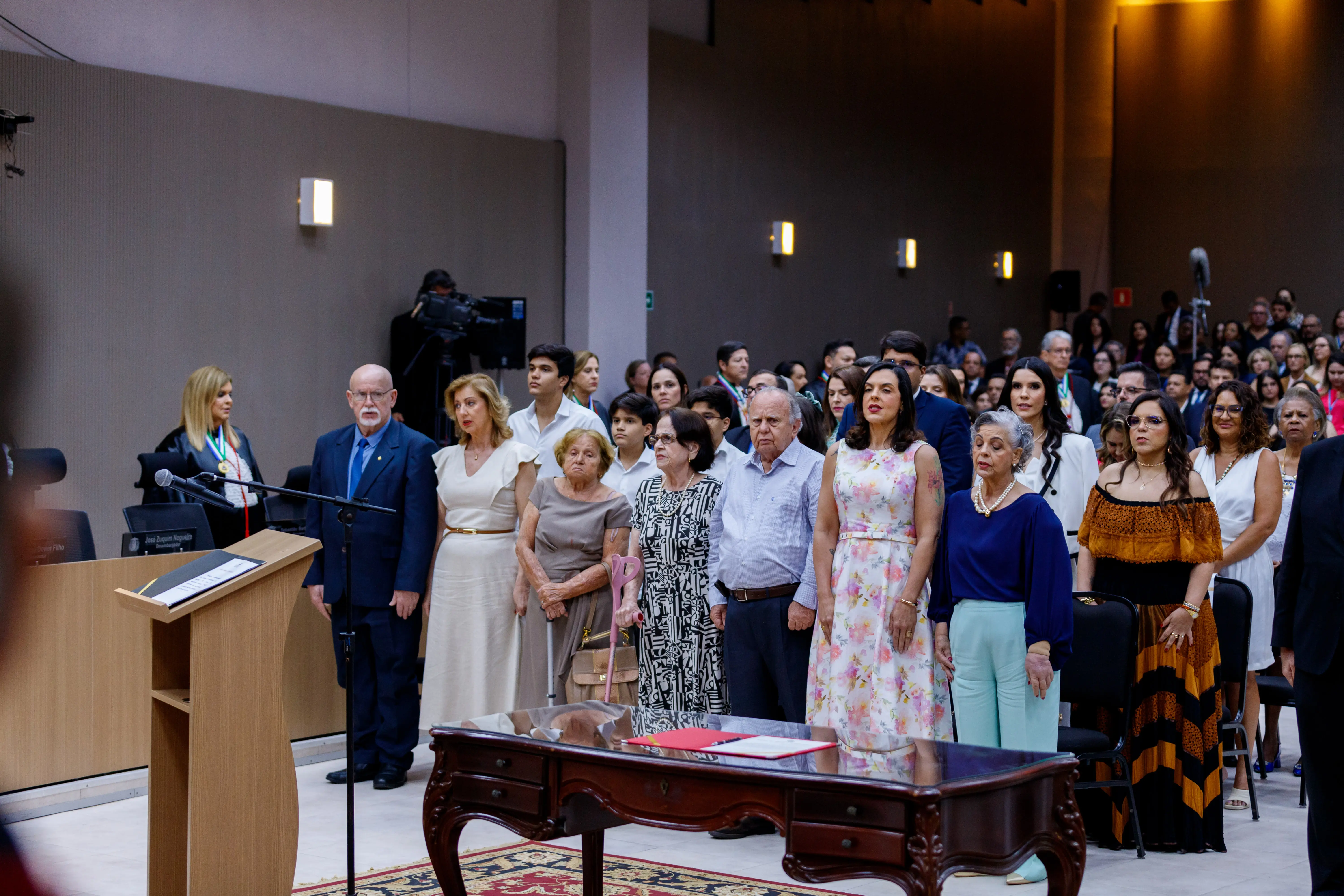 Um grupo de pessoas está reunido no plenário, voltado para a frente. Em primeiro plano, várias mulheres estão em fila, vestidas com roupas formais.