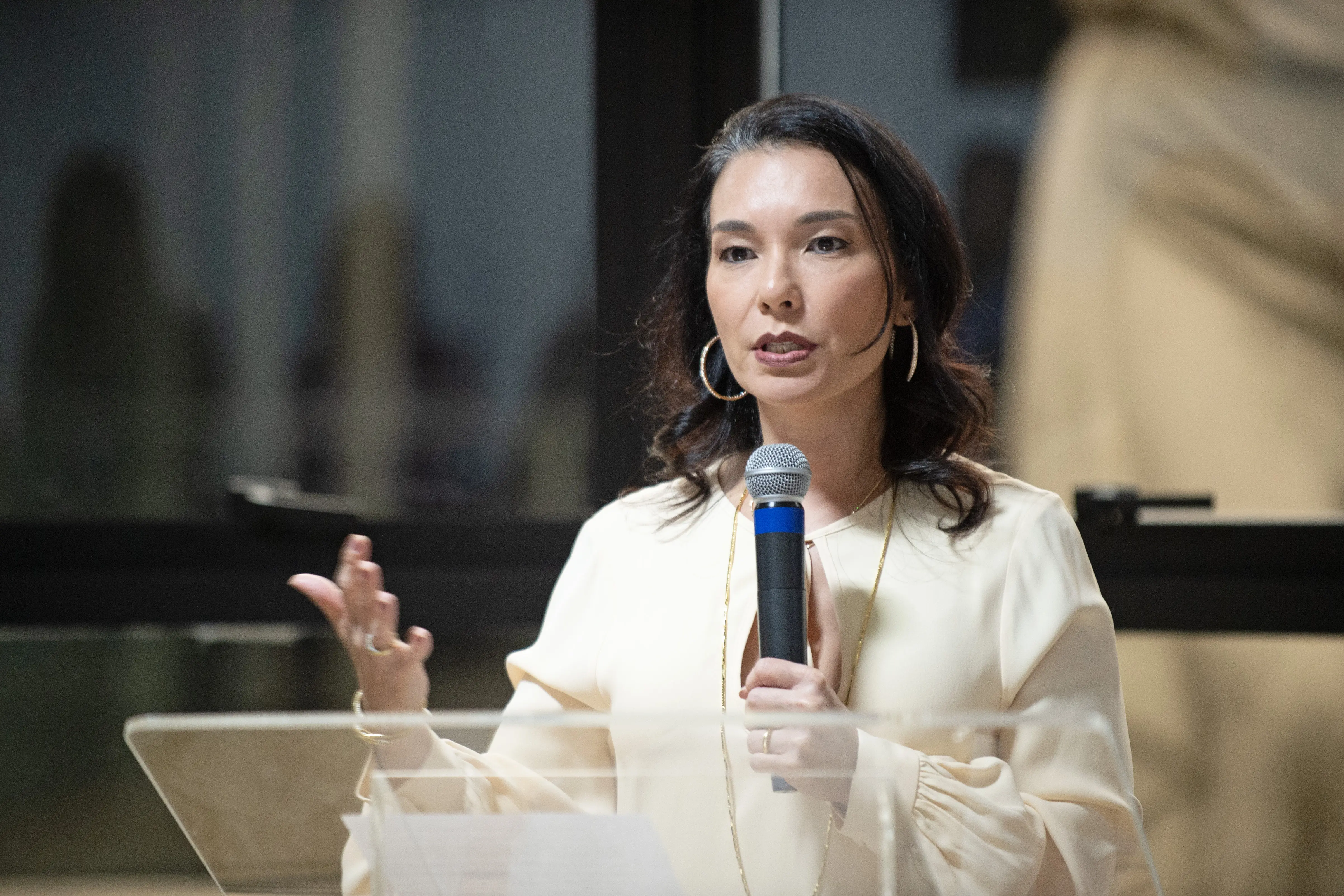 Juíza Hanae Yamamura durante solen idade de 20 anos da estrutura do Fórum de Cuiabá. Ela é uma mulher com traços orientais, branca, de cabelos curtos pretos. Usa um  vestido branco e fala ao microfone