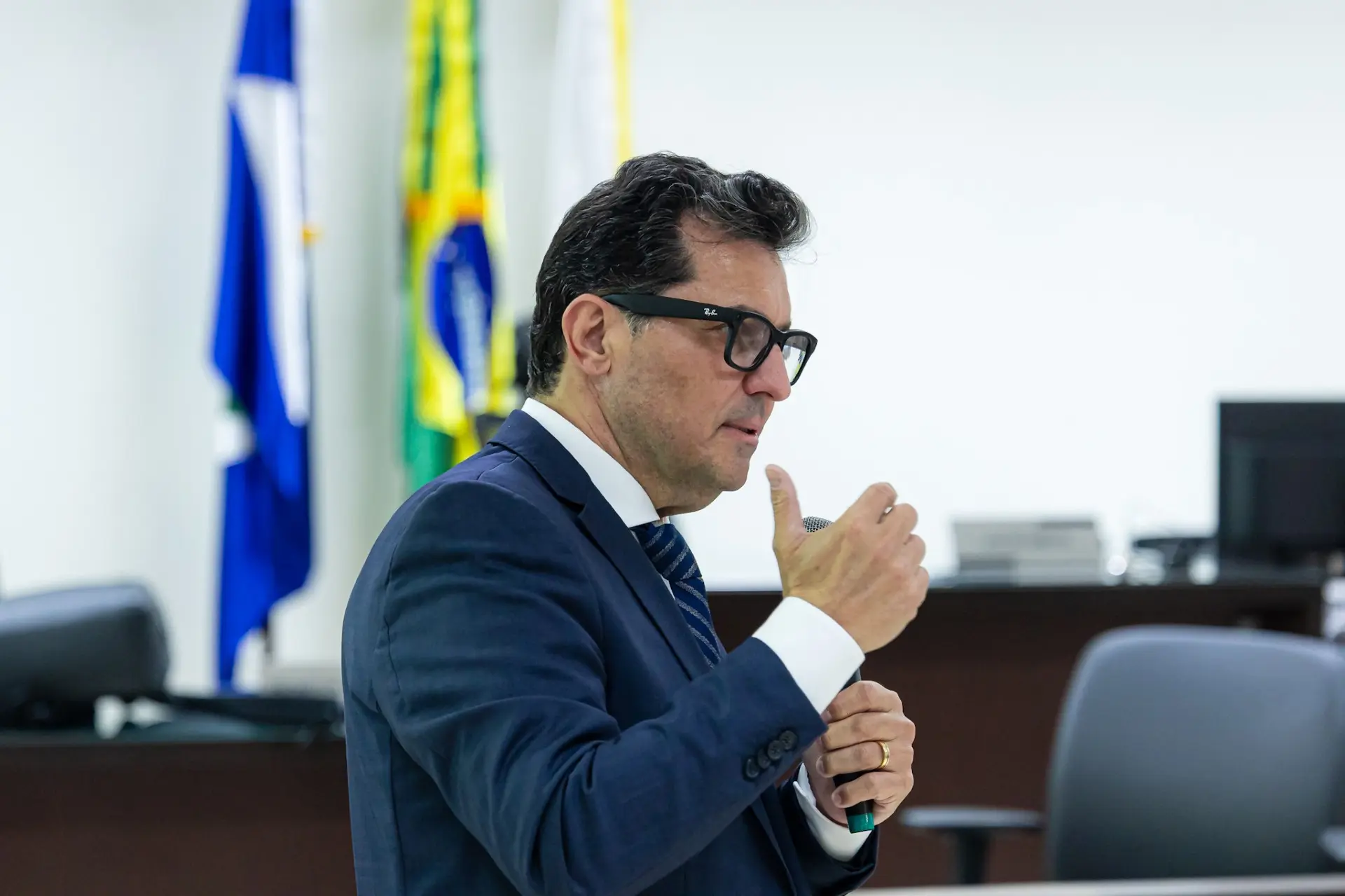 Foto horizontal do juiz Jeverson Luiz Quintieri falando ao microfone. Ele é um homem branco, de cabelo escuro, usando terno e gravata azul marinho, óculos de grau de armação preta. Ao fundo aparecem mastros com bandeiras de Mato Grosso e do Brasil.