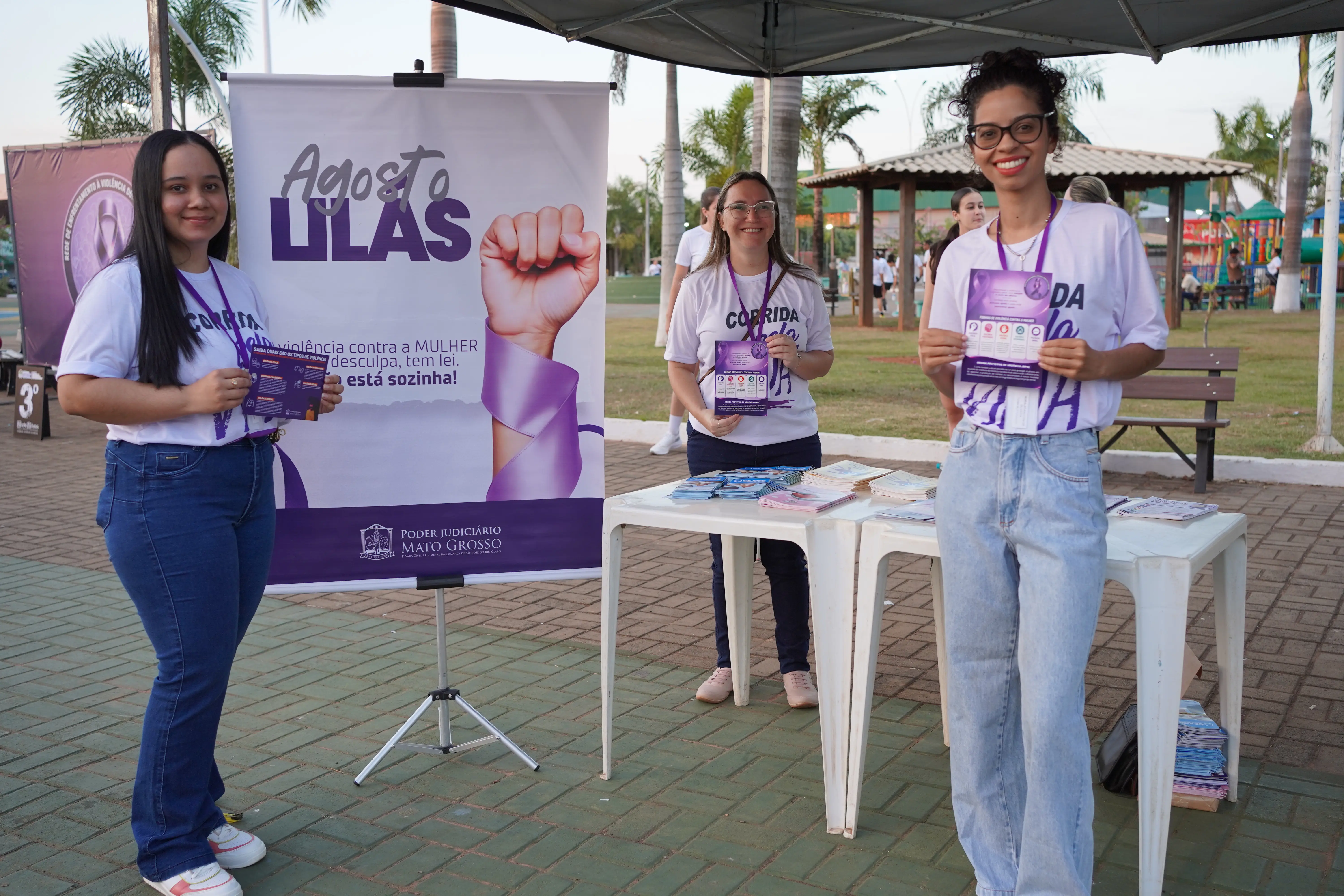 Três mulheres seguram folhetos educativos ao lado de banner sobre a campanha de combate à violência contra a mulher, montado em espaço público arborizado.
