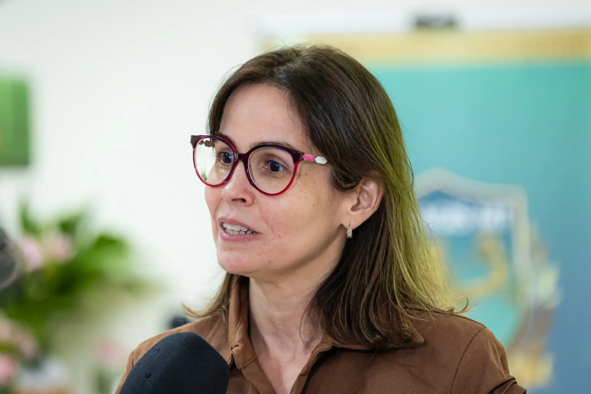 Foto horizontal da juíza Augusta Prutchanski durante entrevista à TV.  Ela é uma mulher branca, de cabelos lisos, castanhos claros, na altura dos ombros, usando óculos de grau com armação vermelha, vestindo camisa marrom de botões. 