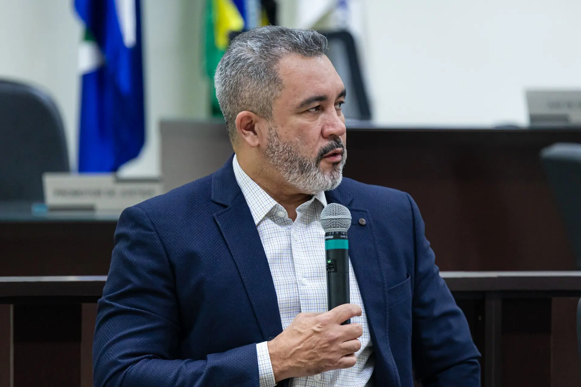 Foto horizontal que mostra o juiz Antonio Peleja falando ao microfone, no plenário do júri de Barra do Garças. Ele é um homem pardo, com cabelos grisalhos curtos e barba cheia, veste blazer azul-marinho e camisa xadrez clara. 