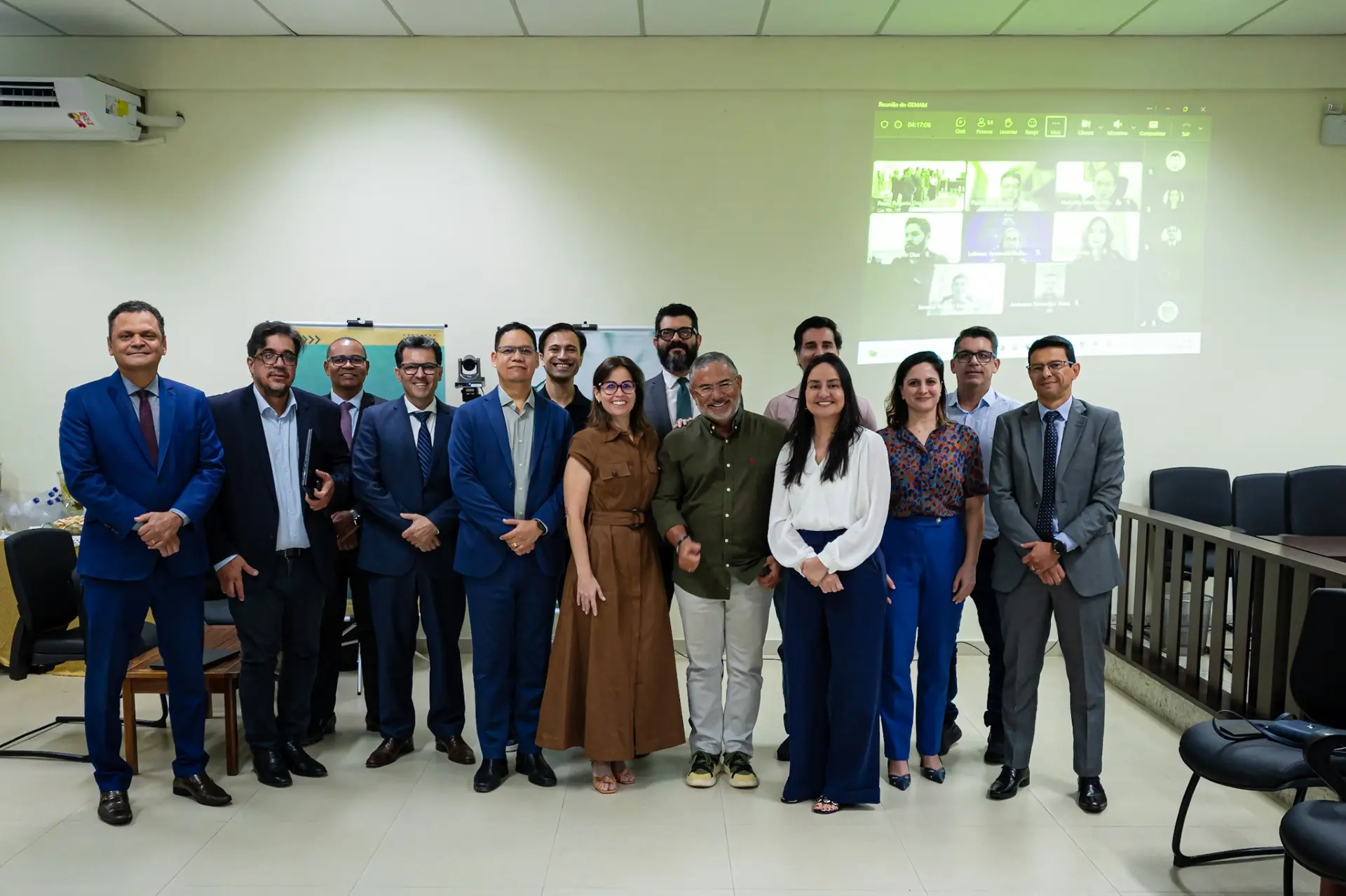 Foto horizontal que mostra um grupo de 15 magistrados e magistradas posando para a foto, em pé, no centro do plenário do tribunal do júri do fórum de Barra do Garças. Ao fundo, no telão, aparecem rostos de outros participantes on-line.