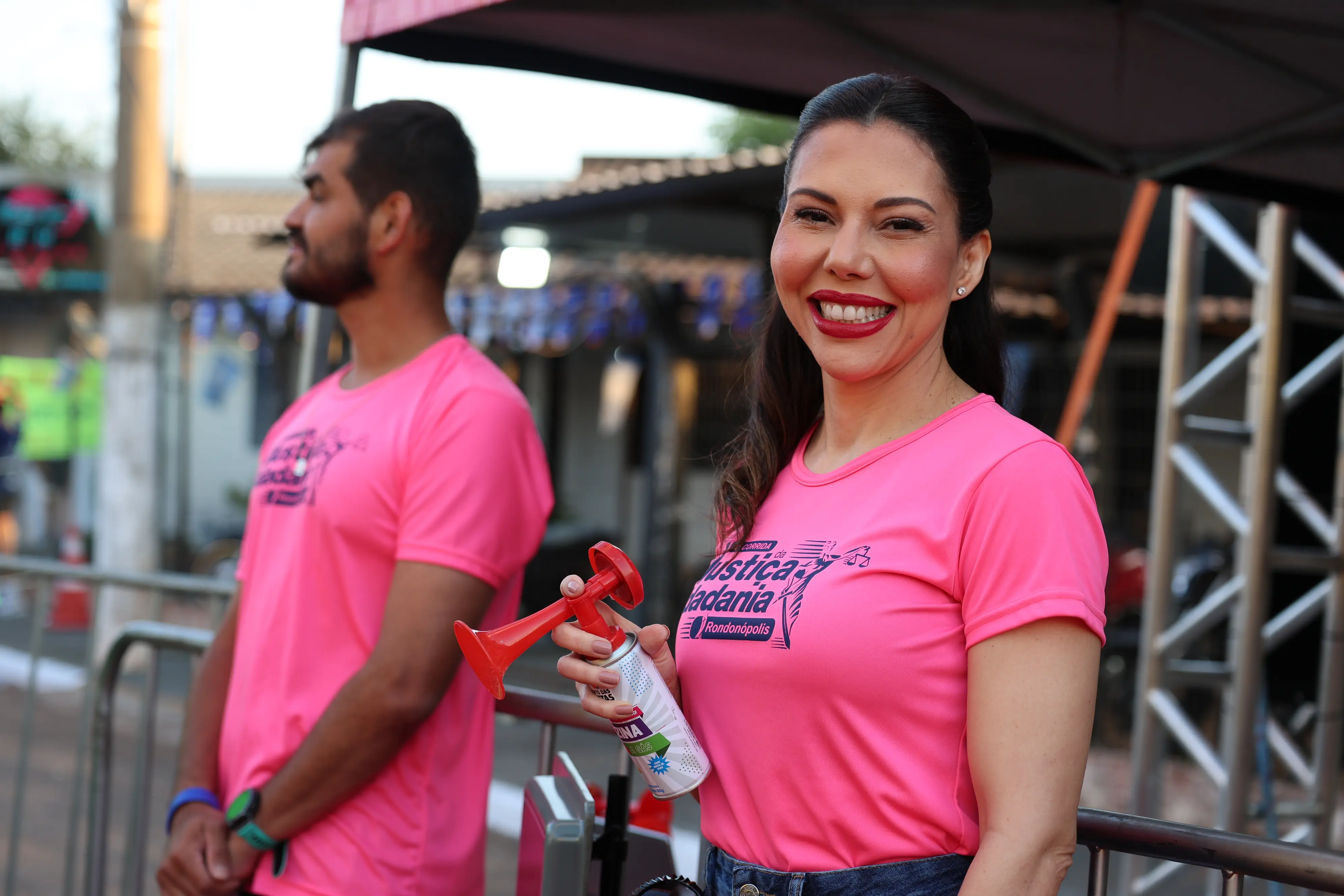 Uma mulher sorri segurando uma corneta de ar, vestindo camiseta rosa alusiva a uma prova esportiva. Ao fundo, outro participante trajado da mesma forma observa o local.