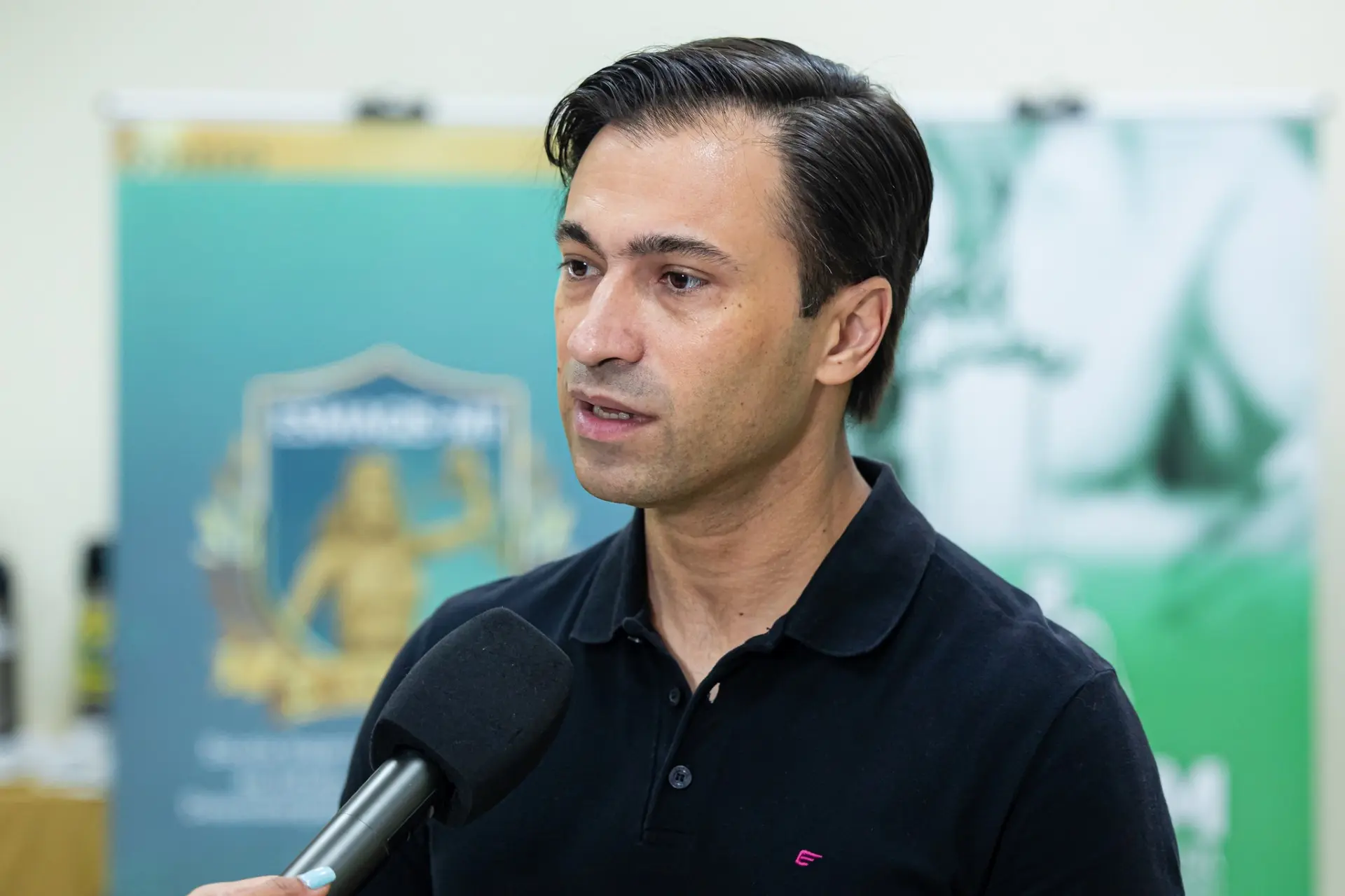 Foto horizontal do juiz Marcelo Bento. Ele é um homem branco, de cabelos escuros e curtos, usando camisa polo preta. Ele fala olhando para o lado perto de um microfone de televisão. Ao fundo, um painel verde com logos e textos desfocados.