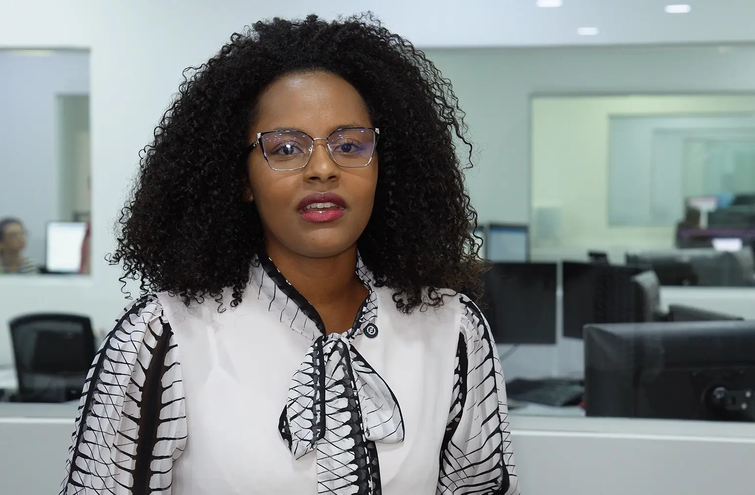 Foto horizontal que mostra a estagiária Mariana Eduarda em pé, olhando para a câmera. Ela é uma jovem negra, de cabelo cacheado, volumoso, preto, solto, na altura dos ombros. Ela usa camisa branca com estampa preta no laço, na gola e nas mangas e óculos de grau. Ao fundo, ambiente de escritório. 