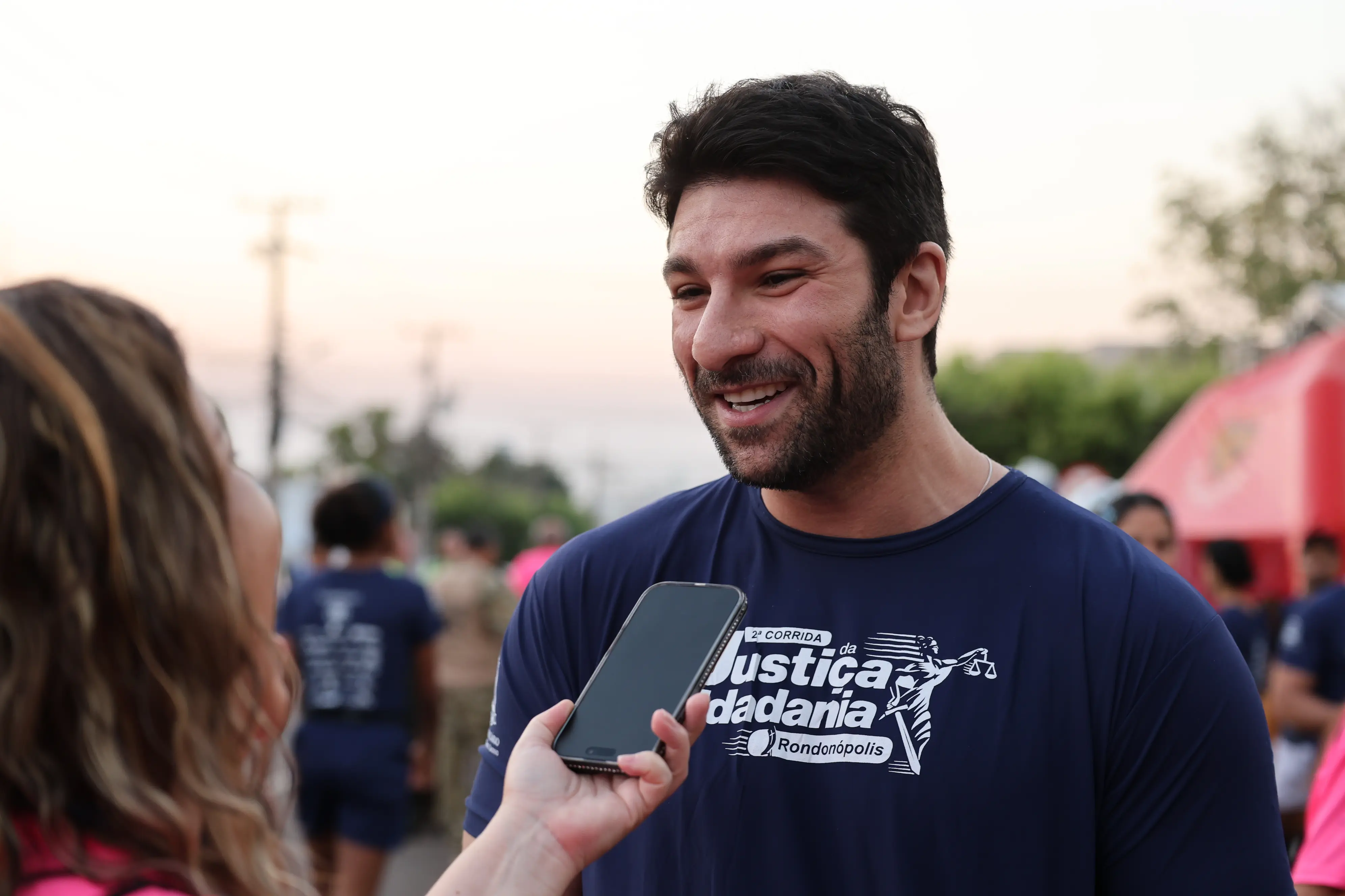 Um homem sorri durante entrevista concedida a uma repórter, que aproxima um celular para captar o áudio. Ao fundo, barracas e outras pessoas compõem o ambiente do evento.