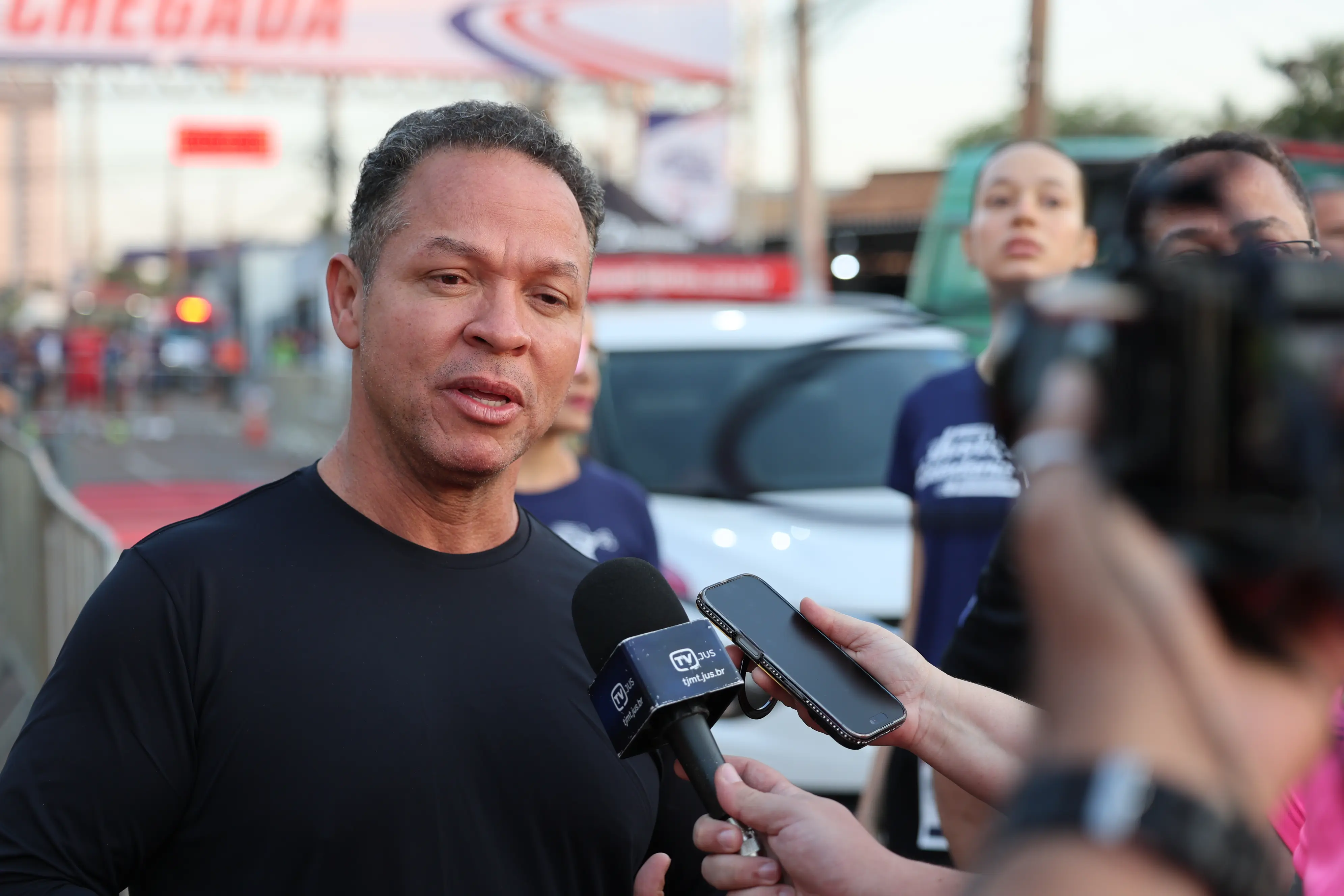 Um homem concede entrevista a microfone com identificação de emissora, durante evento esportivo. Ao fundo, faixa indica a chegada da prova, com veículos e pessoas ao redor.