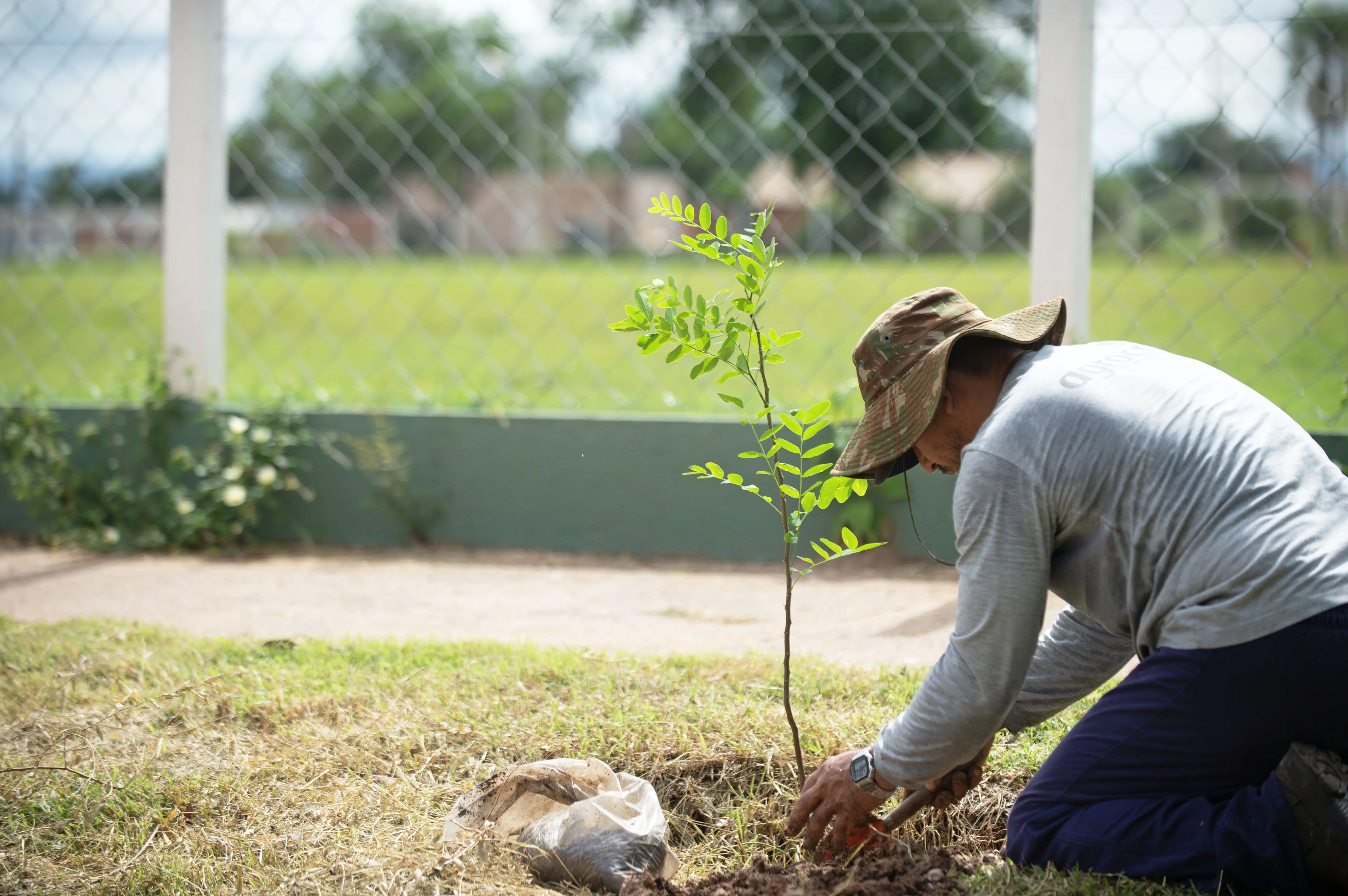 Um jardineiro cuida de uma muda plantada em volta de um campo de futebol. Ele usa blusa de manga comprida e chapéu com estampa camuflada. 