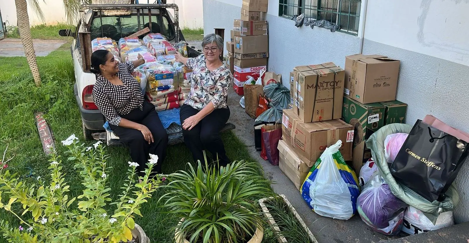 Foto na casa de amparo com as doações arrecadadas pelo TJ e parceiros. Num carro com caçamba estão as doações em sacolas. no chão, várias caixas e duas mulheres sentadas perto das doações estendem as mãos apra mostrar os itens