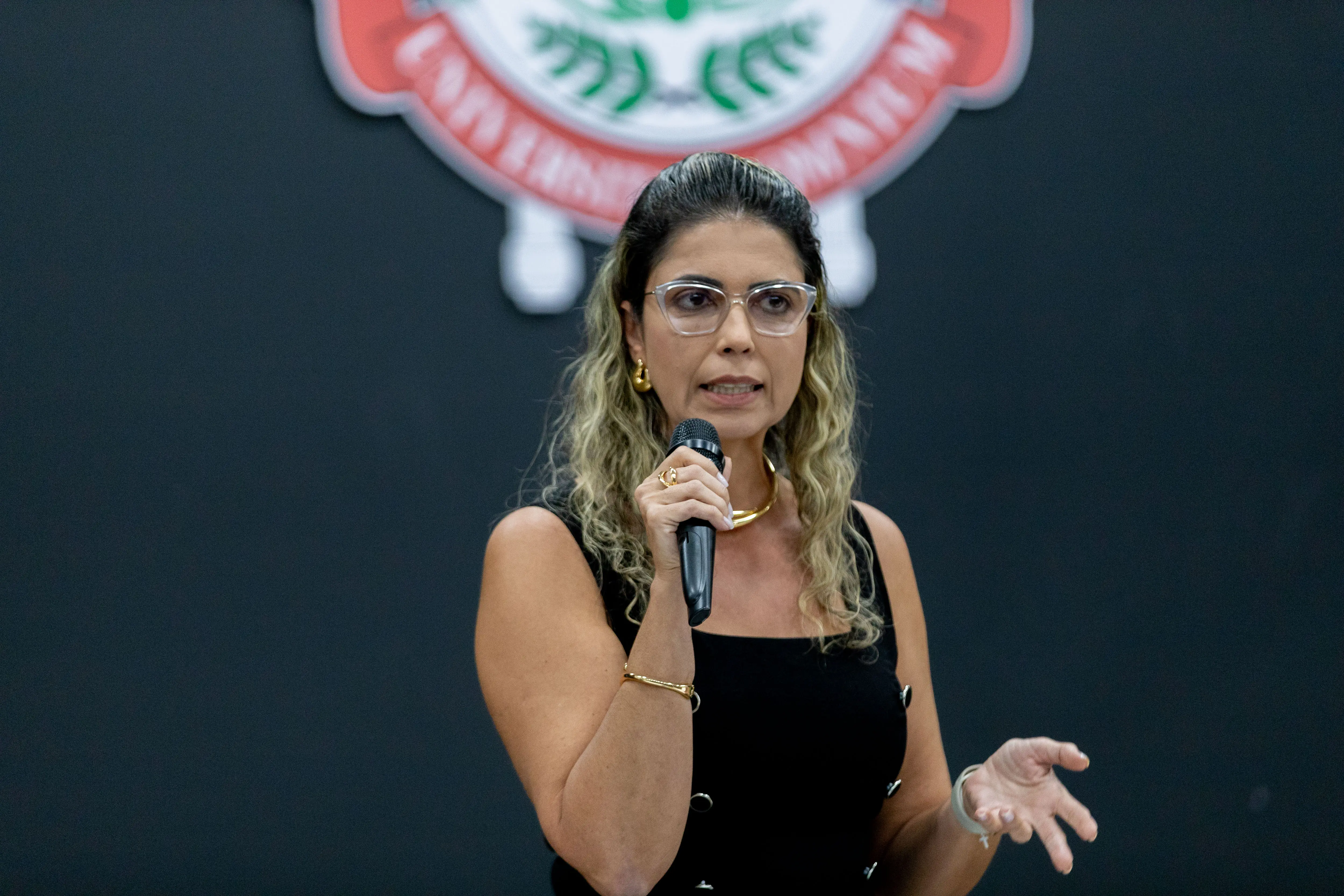 Mulher de cabelos cacheados loiros, com óculos e vestido preto, segura microfone e fala em pé diante de fundo escuro com brasão ao fundo. Ela gesticula com a mão esquerda enquanto apresenta.