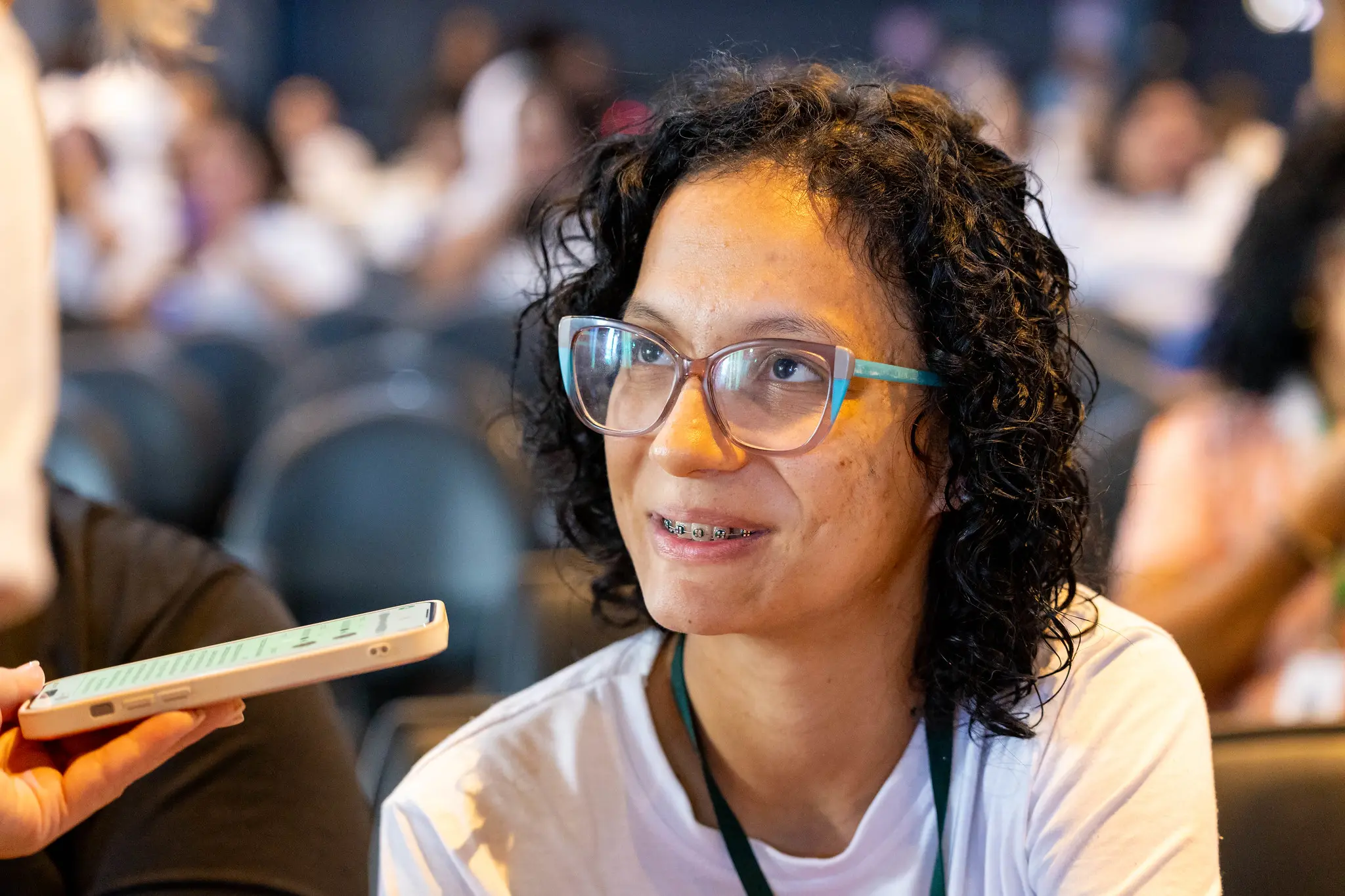 Participante observa com expressão reflexiva durante o evento. Usa óculos de armação clara e camiseta do programa, em ambiente de formação com outros profissionais.