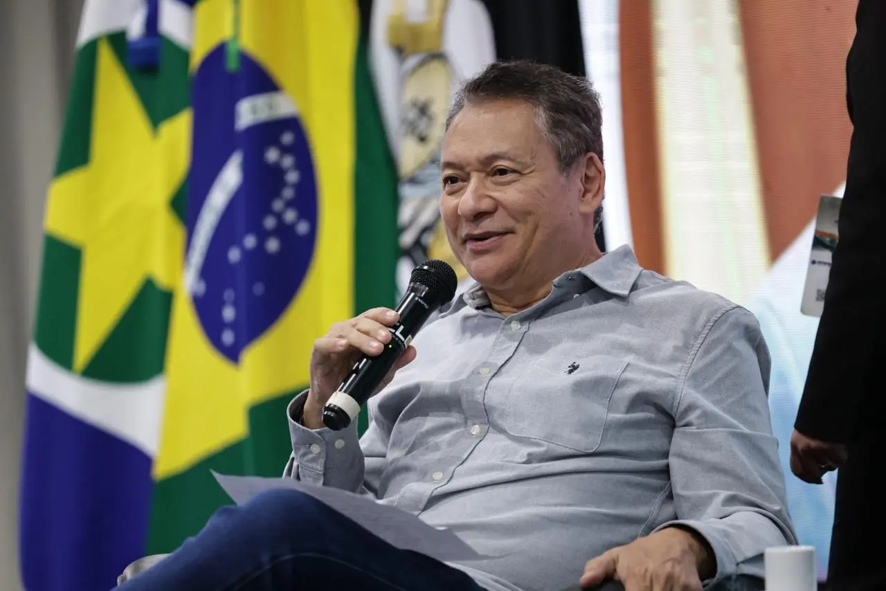 Foto do desembargador Mário Kono sentado em uma poltrona e falando ao microfone. Ele é um senhor magro, pardo, com traços orientais, cabelos grisalhos, usando alça jeans e camisa social cinza.