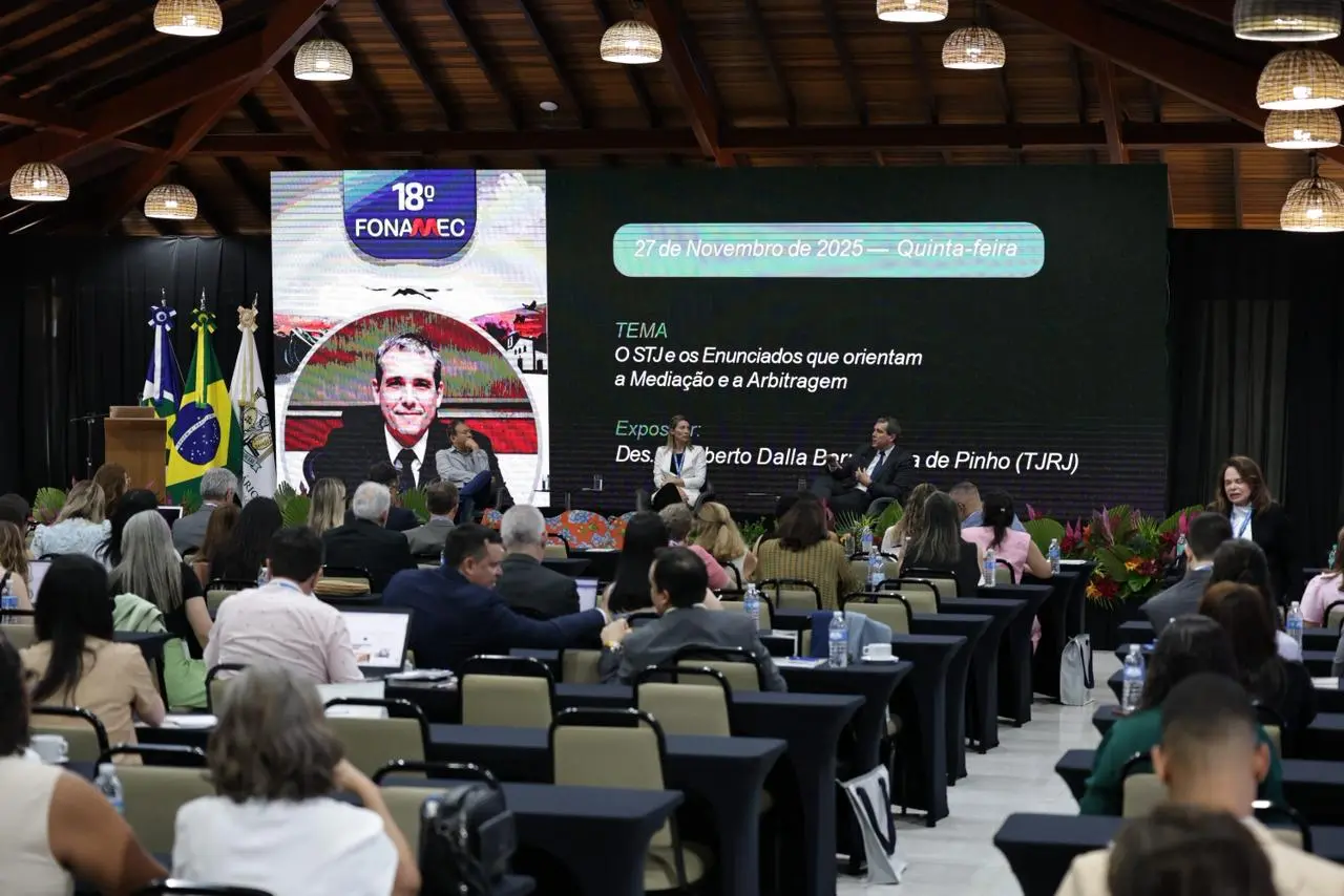 Foto em plano aberto que mostra o auditório lotado de pessoas assistindo a palestras do Fonamec. No palco, estão sentados dois palestrantes e a mediadora e há um telão de LED com a arte do evento.