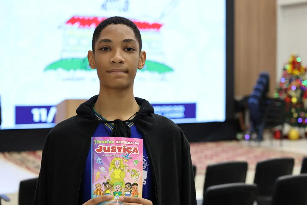Estudante José Mateus Bernardo Grobe da Silva posa sorrindo para a foto, segurando uma cartilha do projeto Justiça e Cidadania. Ele é um adolescente negro, magro, de cabelo preto e curto, usando uniforme azul e casaco preto.