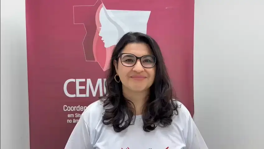 Juíza Lorena Malhado aparece sorrindo, usando óculos e camiseta branca do evento, diante do banner rosa do CEMULHER. O local é interno, bem iluminado, com fundo neutro que destaca sua participação na ação institucional.