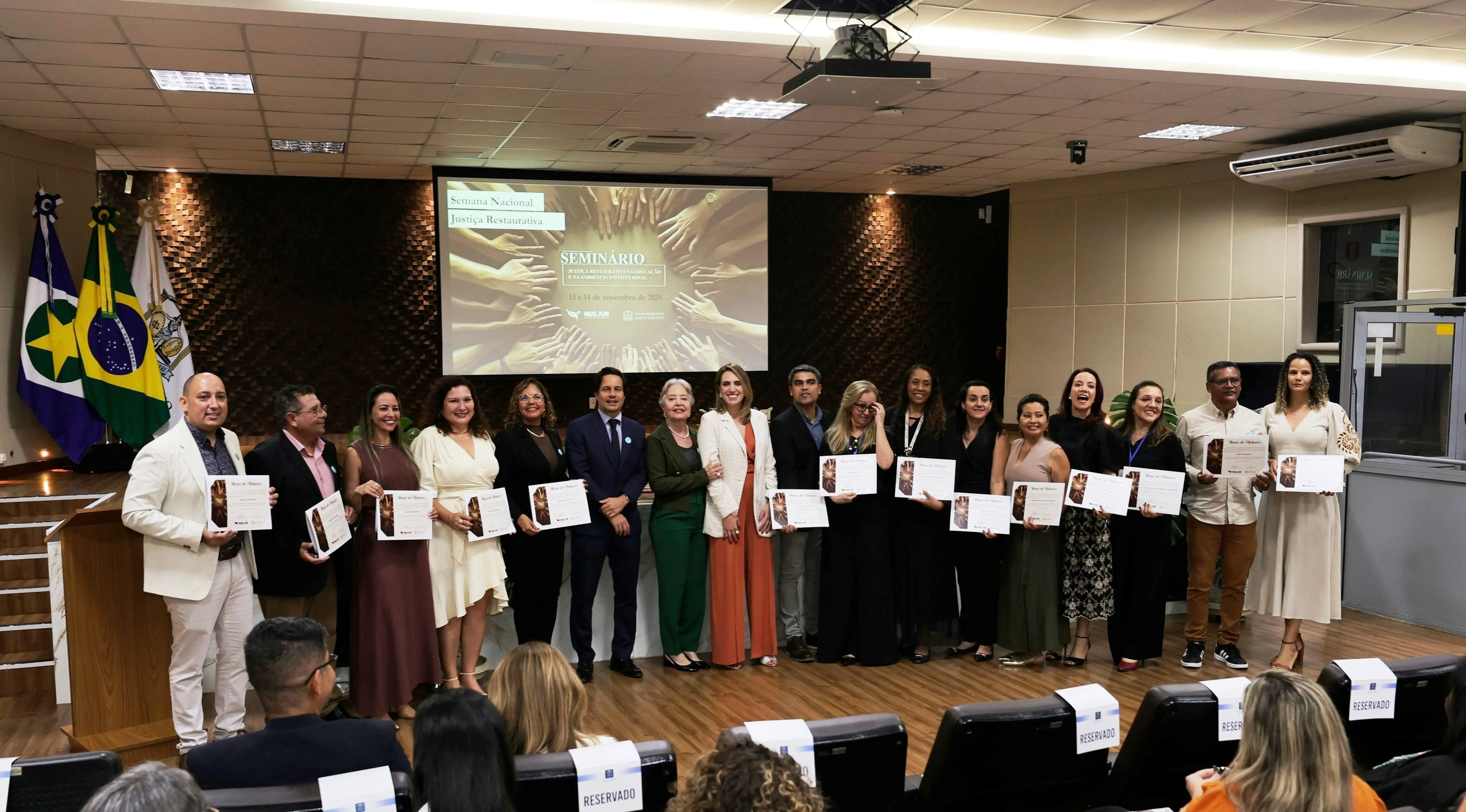 Grupo de cerca de vinte pessoas posadas no palco segurando certificados. Atrás deles, tela exibe "SEMINÁRIO". Plateia sentada observa. Bandeiras do Brasil e outra ao fundo. Ambiente formal com iluminação profissional.