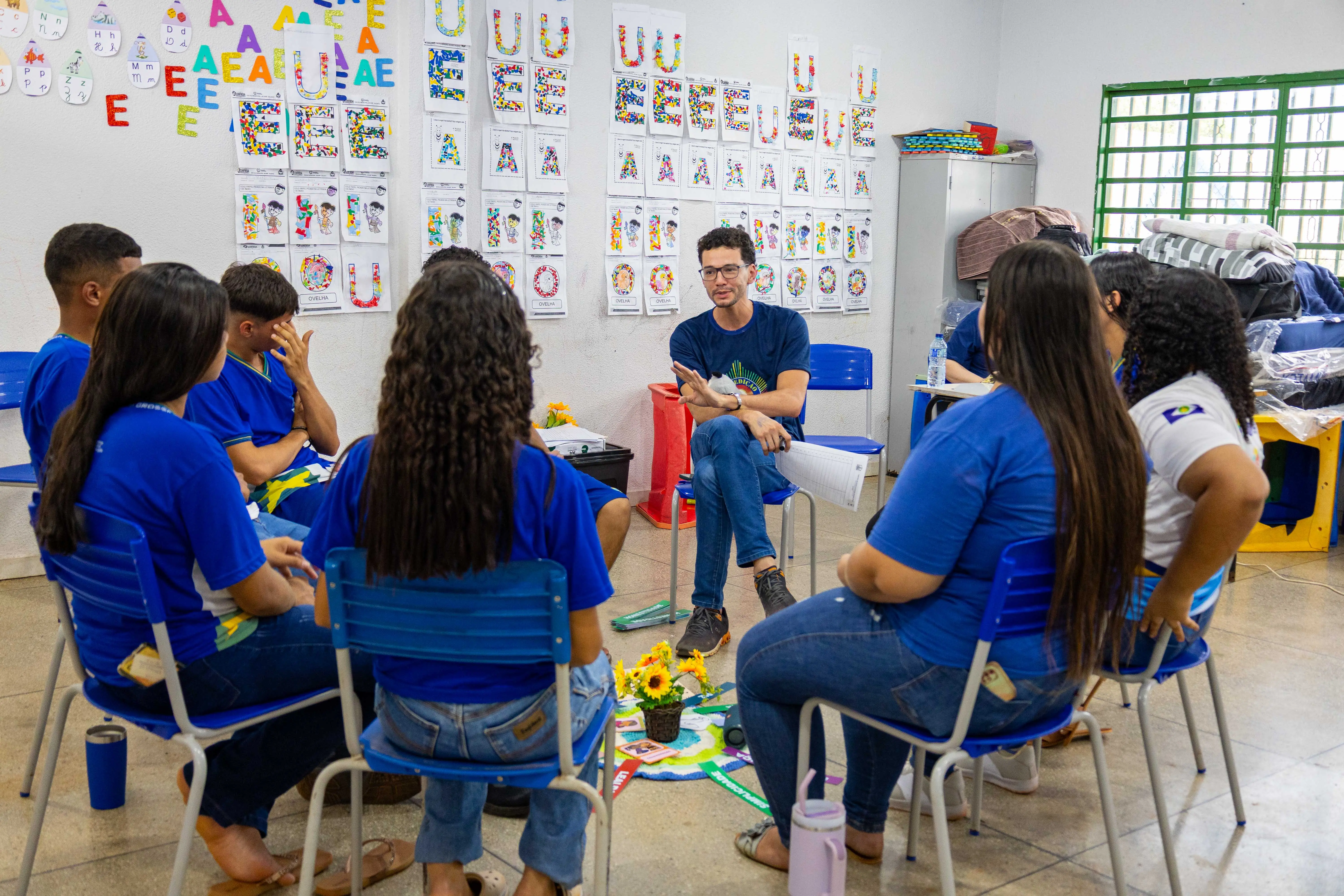 Em sala escolar decorada com letras e atividades infantis nas paredes, um mediador conduz roda de conversa com jovens sentados em círculo. Eles usam camisetas azuis e participam atentamente, em ambiente simples e acolhedor.