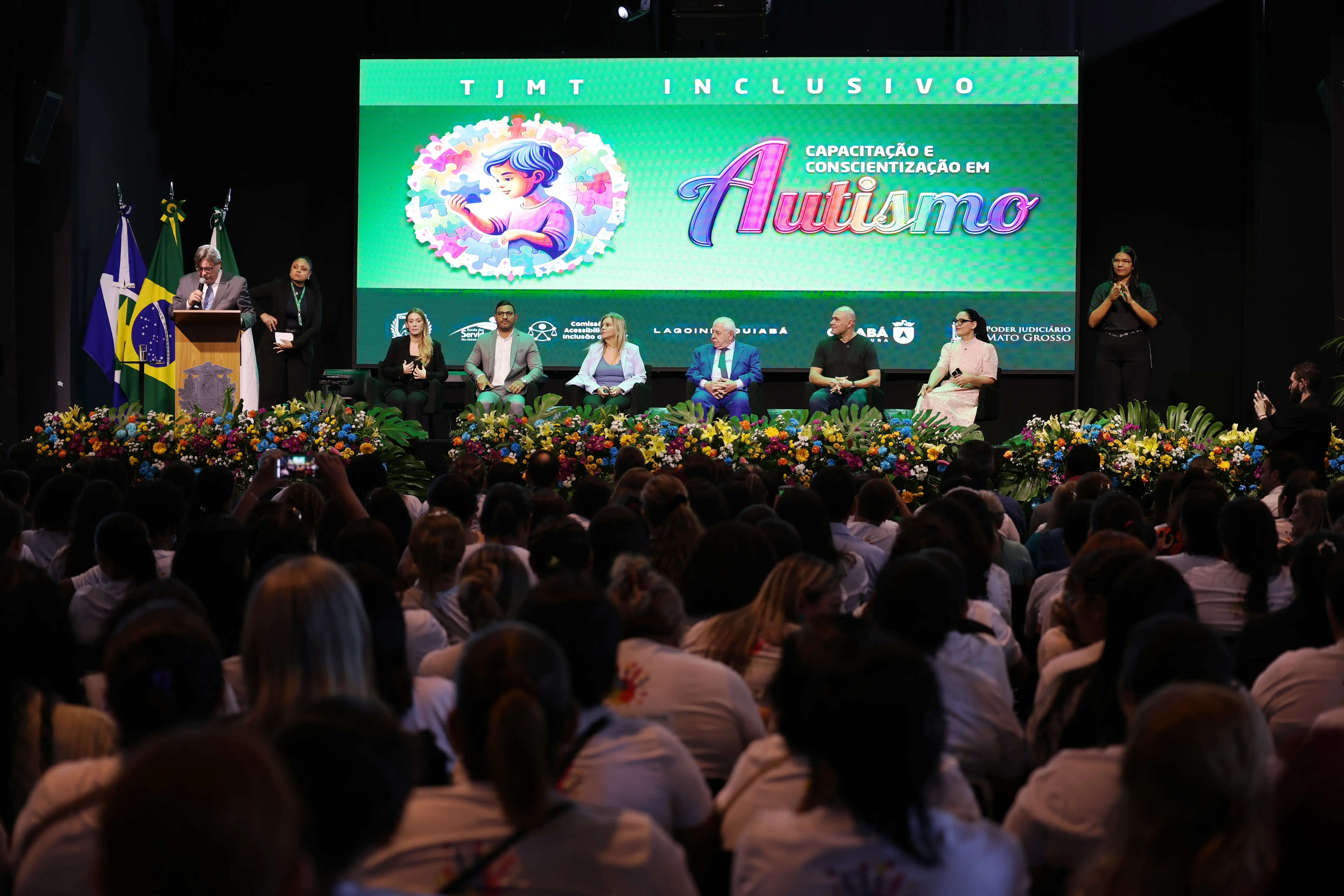 foto em plano aberto que mostra o evento TJMT Inclusivo. É possível ver o palco com as autoridades, o público sentado e o telão com a logomarca do evento.