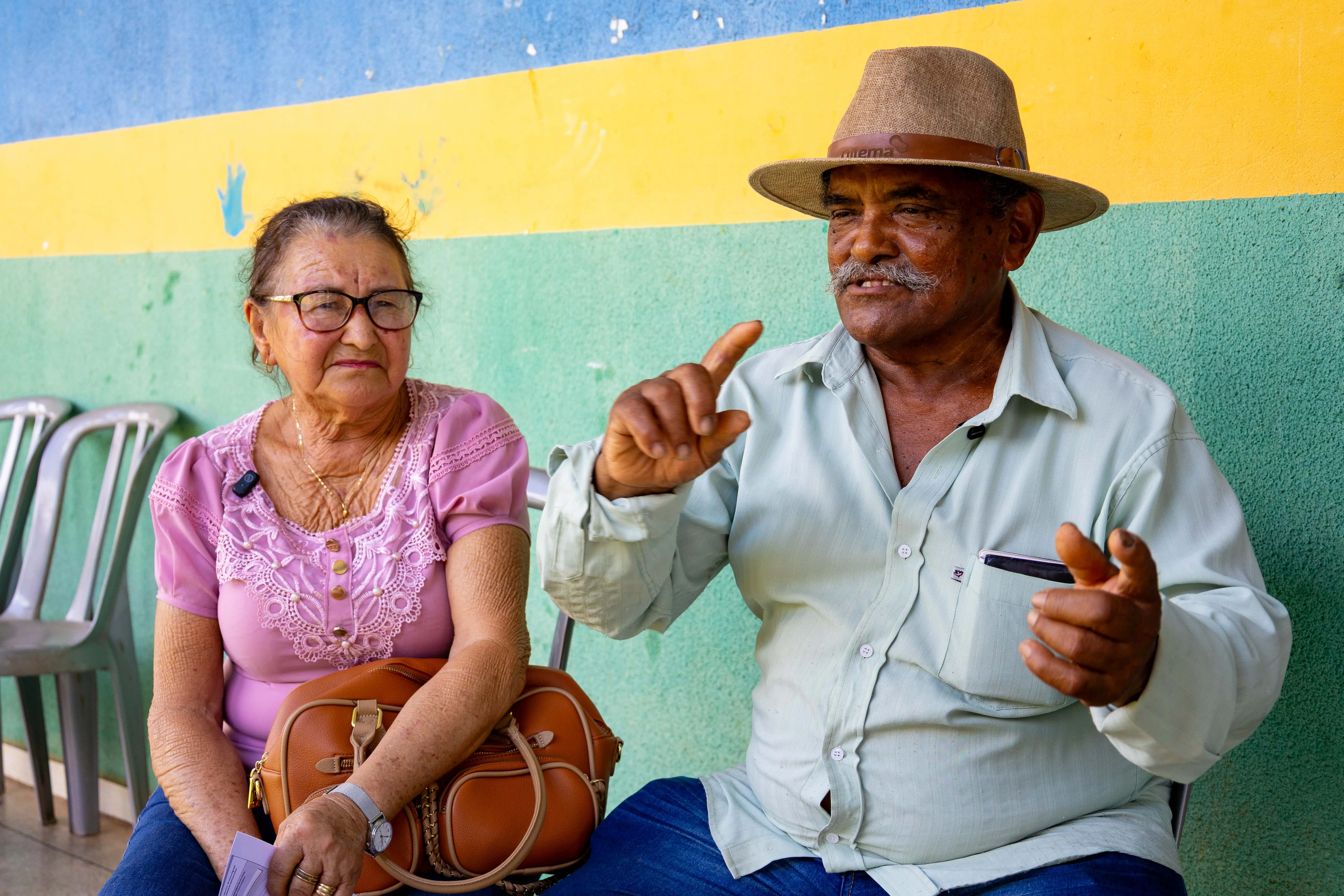 Casal de idosos sentado em banco diante de parede colorida em azul, amarelo e verde. A mulher, de blusa rosa e óculos, segura bolsa no colo. O homem, de chapéu e bigode, fala gesticulando com entusiasmo.