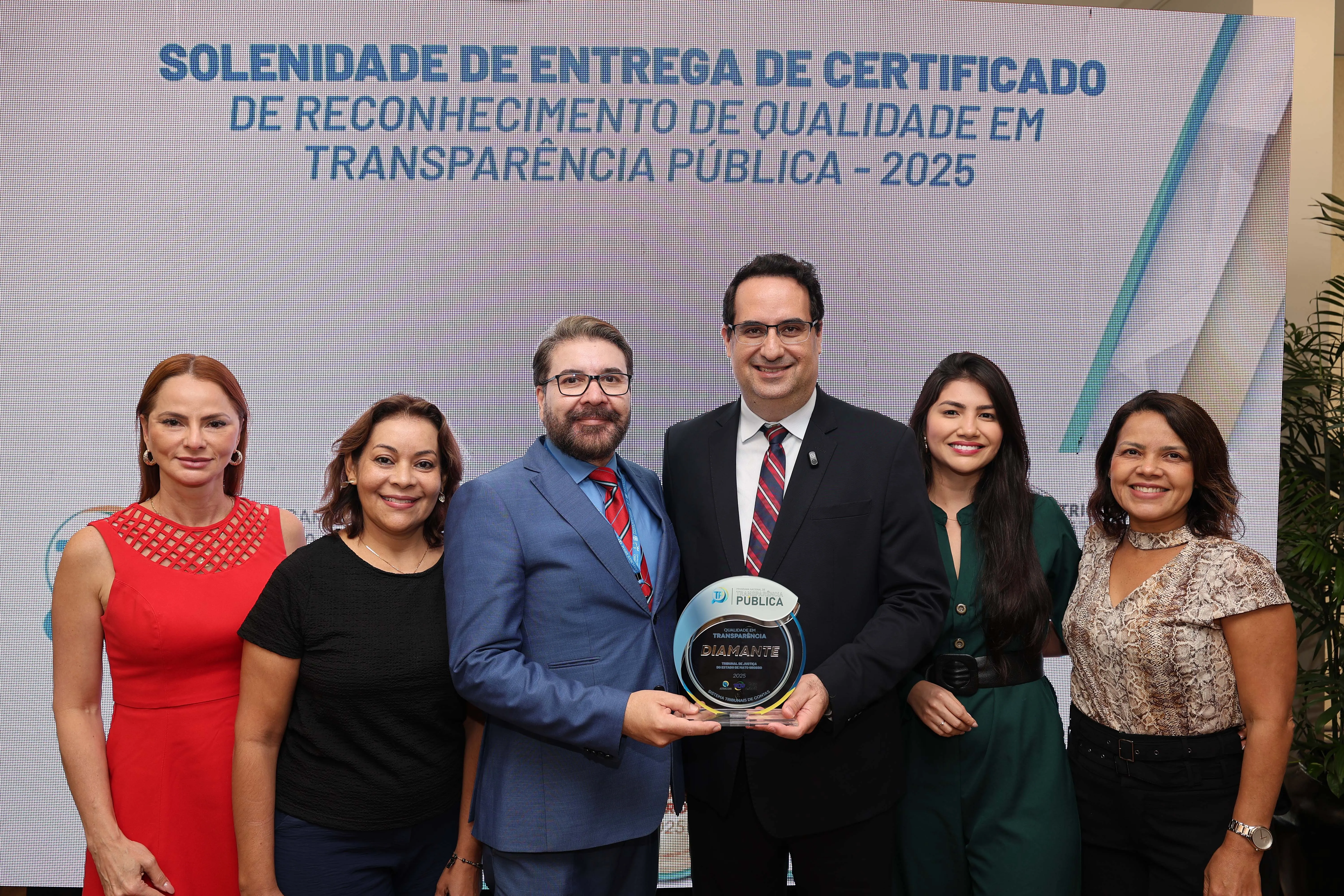 Grupo posa diante do painel da solenidade de certificação em transparência pública. O centro destaca dois homens segurando o troféu Diamante, cercados por mulheres sorridentes, em ambiente iluminado e de celebração institucional.