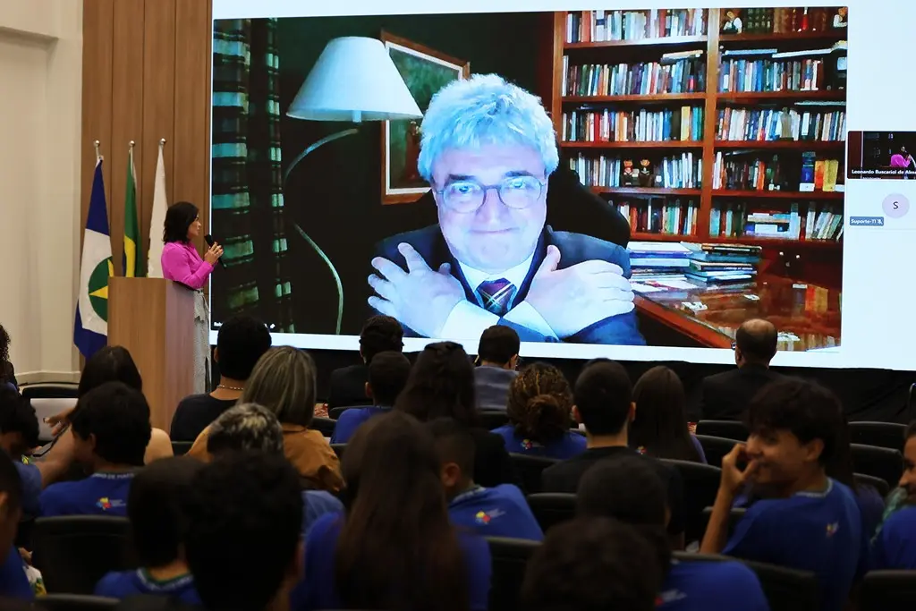 Foto em plano aberto que mostra a palestra remota do desembargador Roberto Bacellar, que aparece no telão sorrindo. Na plateia estão estudantes sentados, de costas na foto.