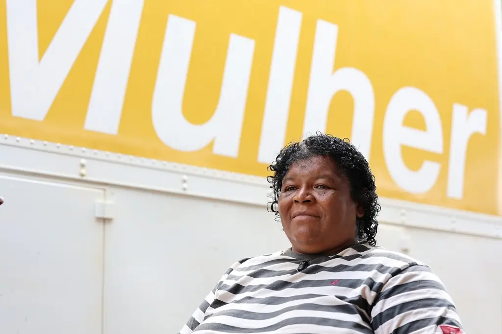 Foto da Maria dos Santos em frente à carreta Sesc Saúde da Mulher. ela é uma mulher de mais de 40 anos, negra, de cabelos pretos, cacheados e curtos, usando uma camiseta listrada preta e branca. 