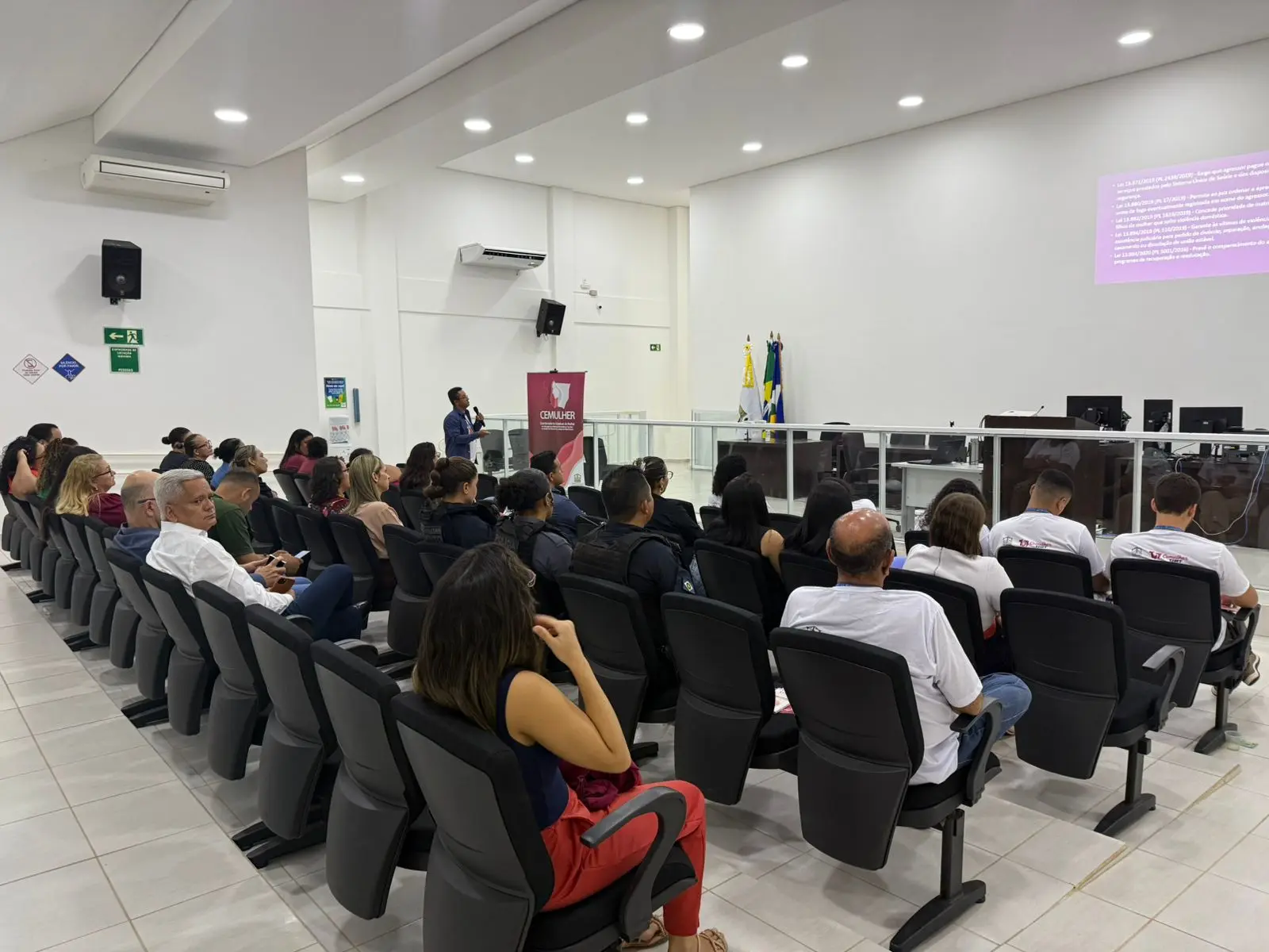 A foto mostra auditório amplo e iluminado, com pessoas sentadas assistindo à apresentação. Um palestrante fala próximo ao banner do CEMULHER. Bandeiras e telas de projeção compõem o ambiente oficial do fórum da comarca.