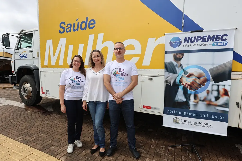 Foto da juíza Cristiane Padim e dos servidores do Nupemec, Vanessa Sabrina e Sebastião José posando para a foto, sorrindo, em frente à carreta Sesc Saúde da Mulher e do banner do Nupemec.