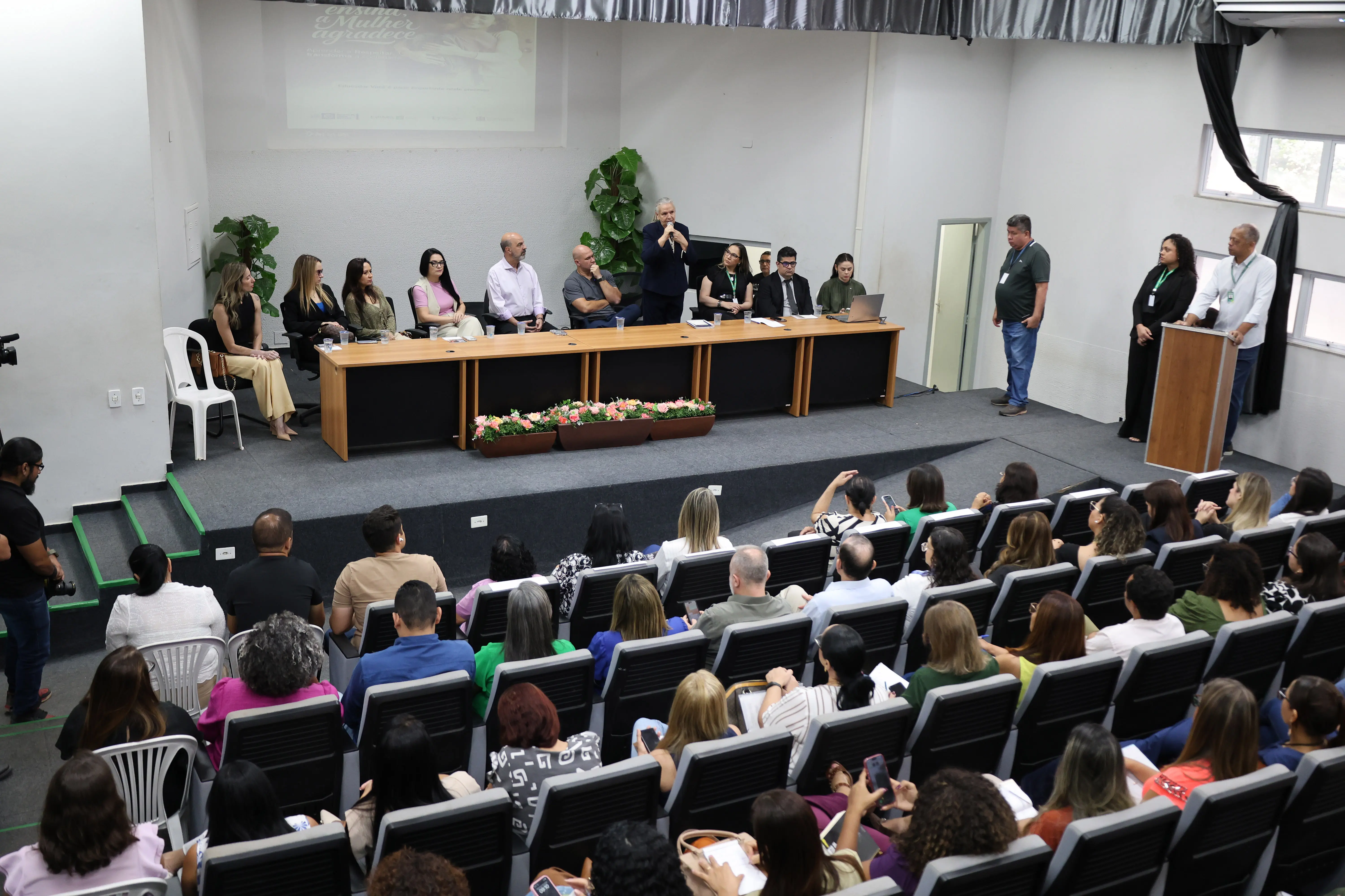 Foto em plano aberto, tirada de cima, que mostra o auditório da Secretaria de Educação de Cuiabá lotada. No placo, sentados no dispositivos de honra estão diversas autoridades do Poder Judiciário, bem como do Executivo e Legislativo de Cuiabá.