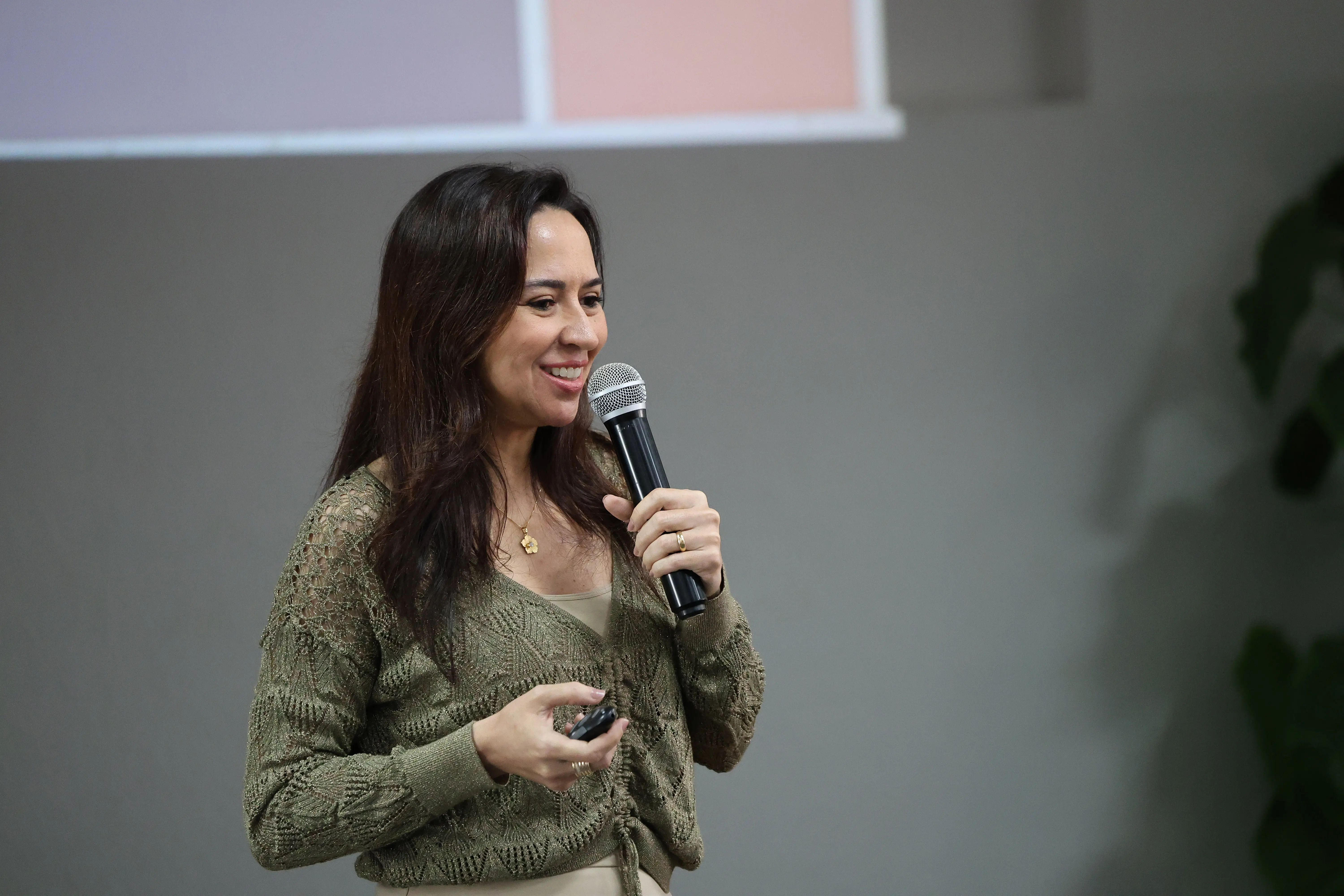 Juíza Tatyana Lopes de Araújo Borges fala sorrindo ao microfone. Ela é uma mulher branca, de cabelos e olhos castanhos, usando vestido bege e moletom de tricô verde e colar dourado com pingente de flor. 
