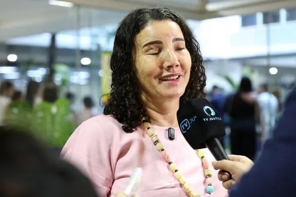 Mulher de cabelos cacheados e blusa rosa dá entrevista sorrindo para a TV Justiça, segurando um microfone. Ao fundo, pessoas circulam em um ambiente interno iluminado e com plantas decorativas.