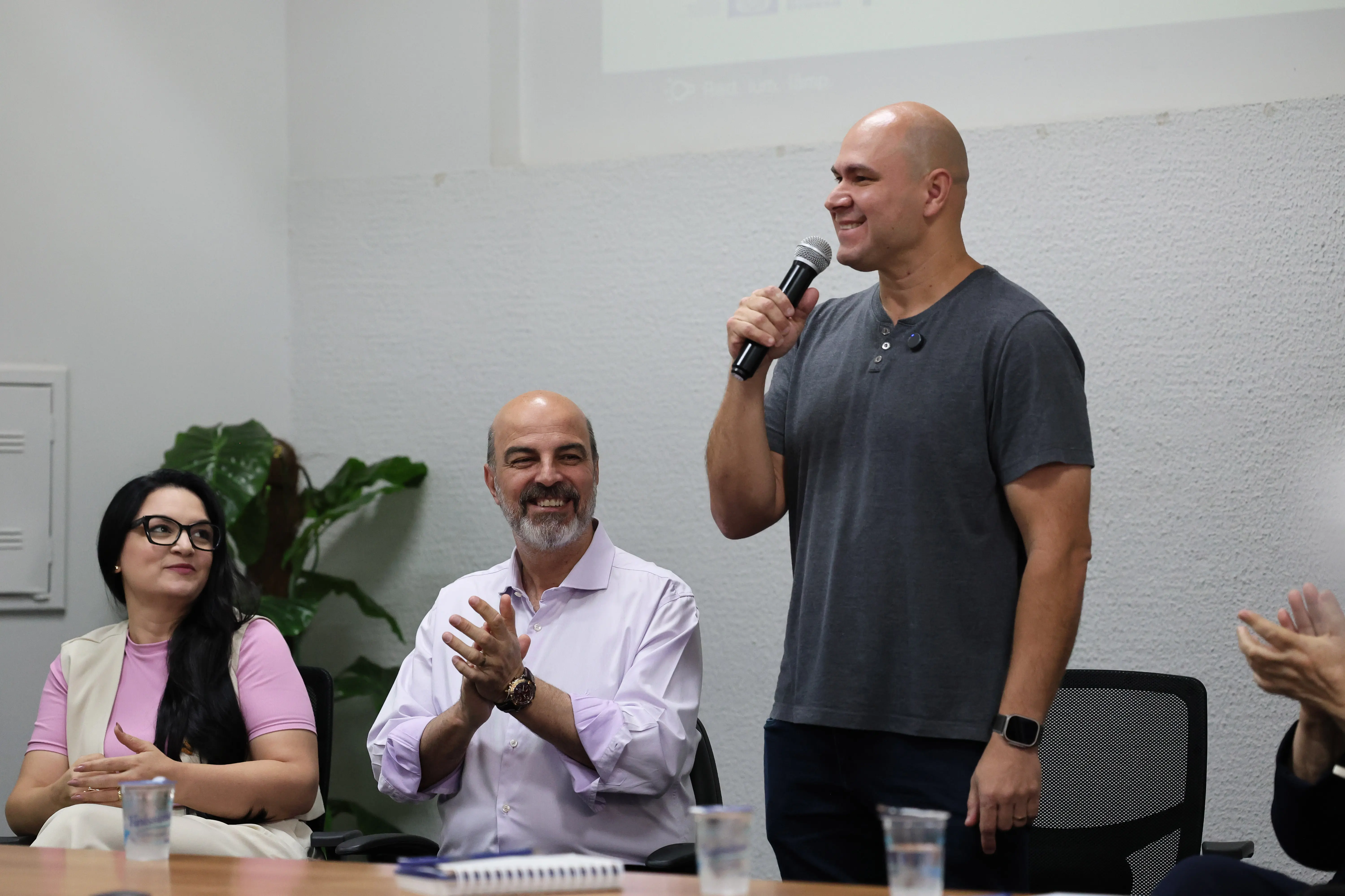 Prefeito Abílio Brunini em pé, falando ao microfone e sorrindo. Ele é um homem branco, careca, usando calça escura e camiseta cinza. Do lado esquerdo da foto, sentados e sorrindo, estão a primeira-dama e vereadora Samantha Íris e o secretário de Educação de Cuiabá, Amauri Fernandes.