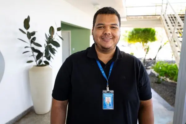 Homem jovem com crachá azul e camiseta preta sorri para a câmera em corredor iluminado. Atrás dele, vasos com plantas e uma escada metálica compõem o ambiente moderno e bem iluminado.