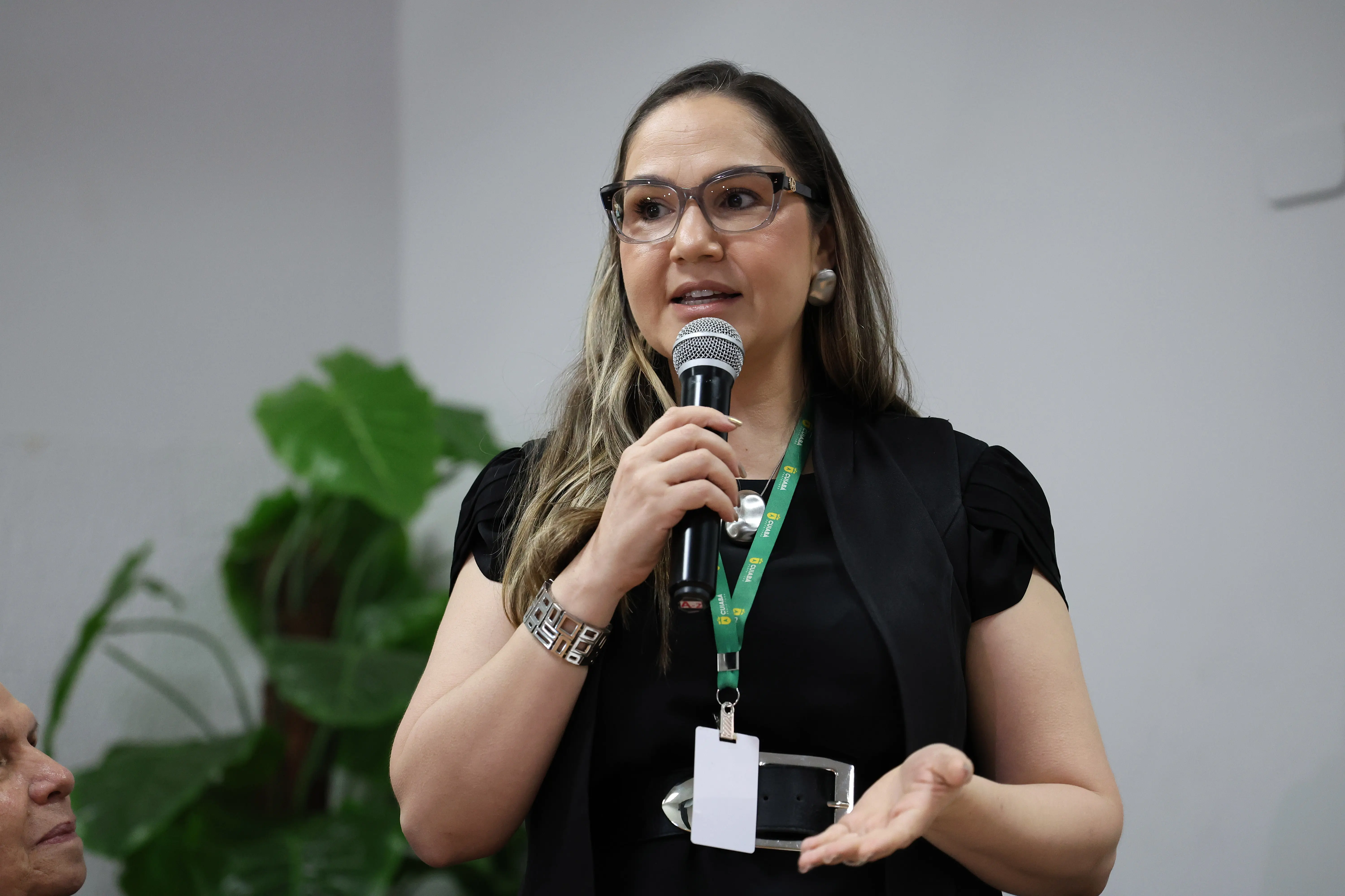 Secretária da Mulher de Cuiabá, Hadassah Suzannah, fala ao microfone. Ela é uma mulher branca, de olhos castanhos claros, cabelos castanhos claros com mechas loiras, usando blusa, colete e cinto pretos, acessórios prateados e óculos de grau.