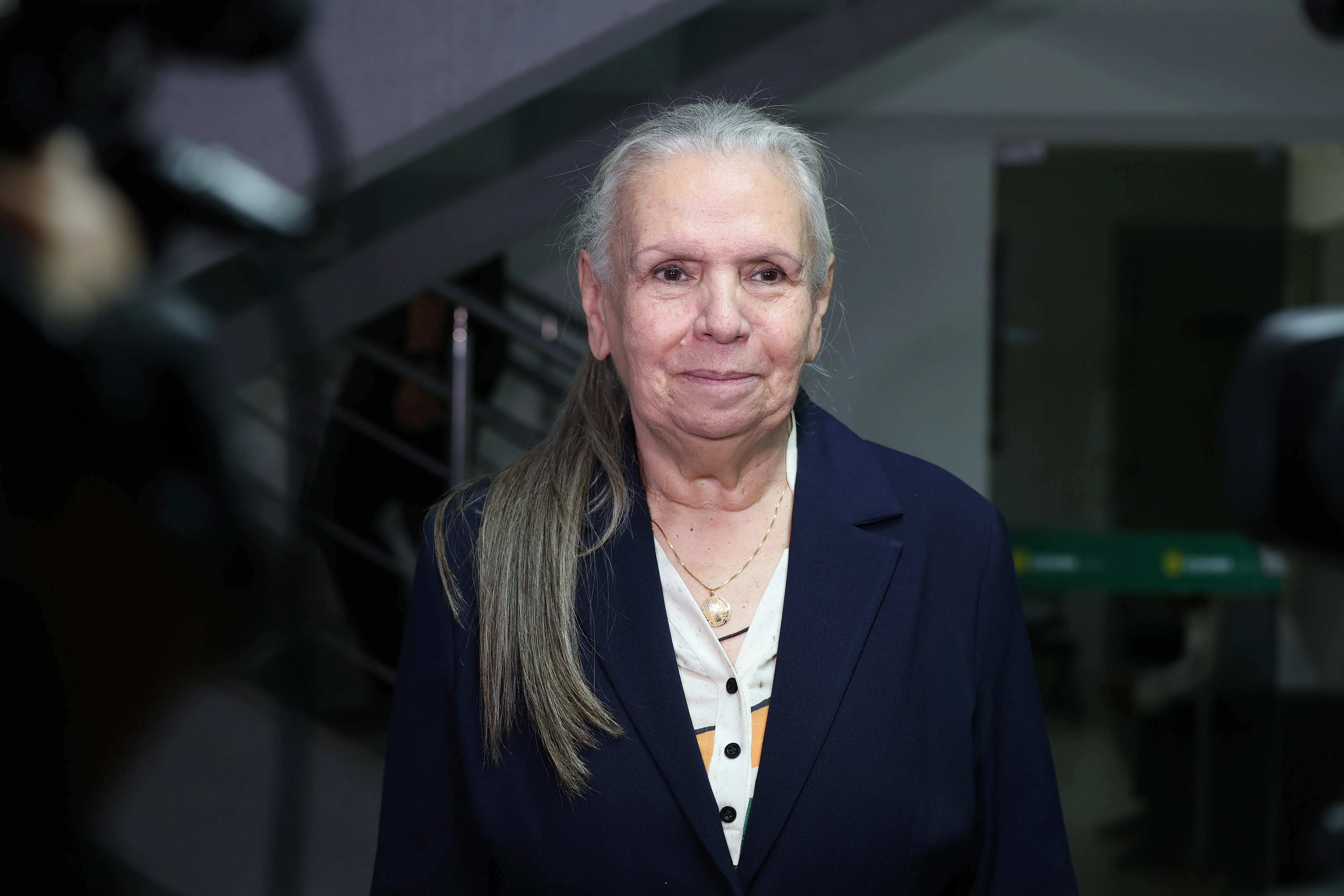  Desembargadora Maria Erotides Kneip em pé e sorrindo. Ela é uma senhora branca, de cabelos grisalhos, usando blazer azul marinho e colar com pingente de Nossa Senhora. 